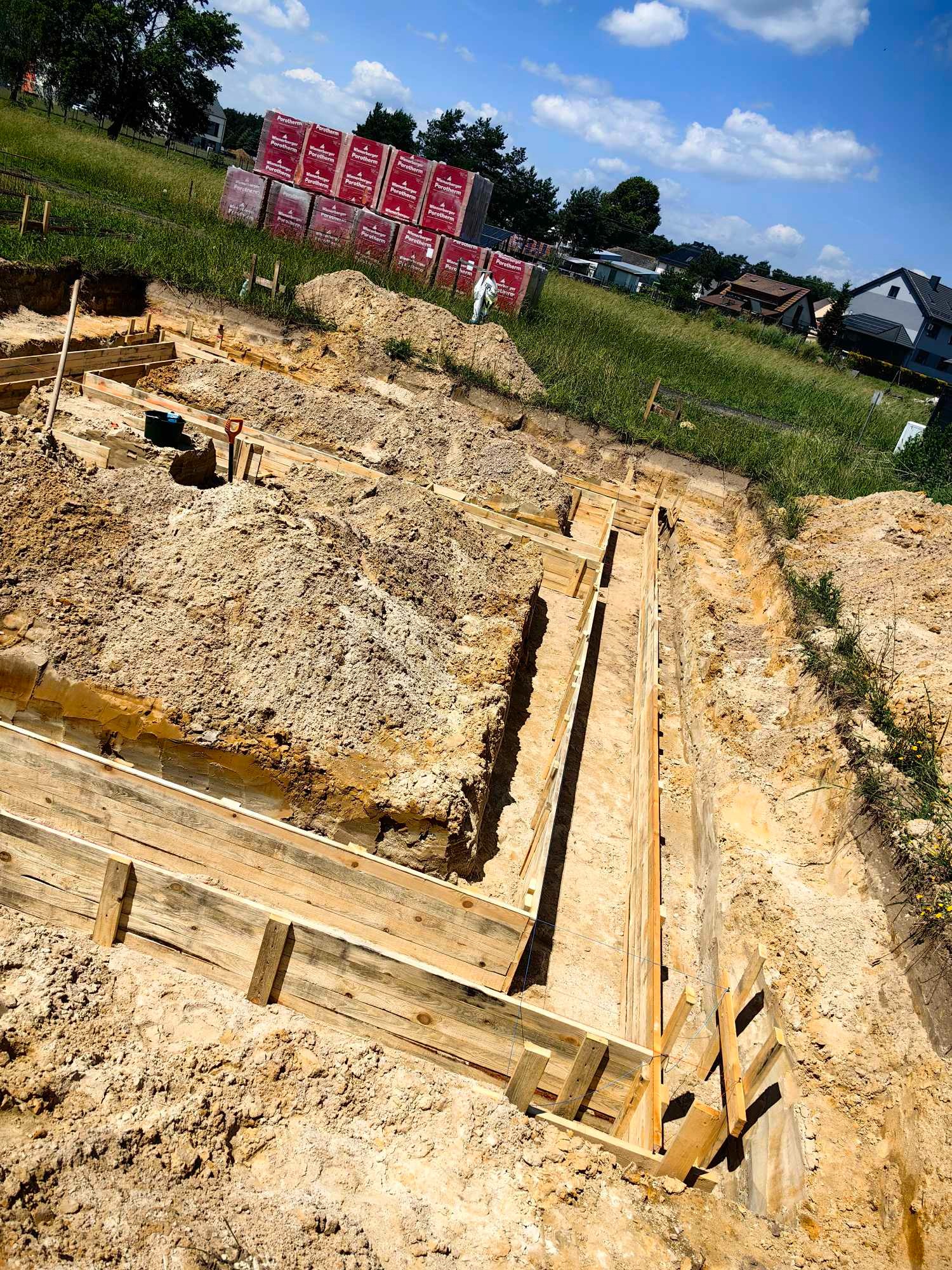 Przygotowanie wykopu pod fundamenty z drewnianym szalunkiem, w tle palety z bloczkami Porotherm i zabudowa jednorodzinna.