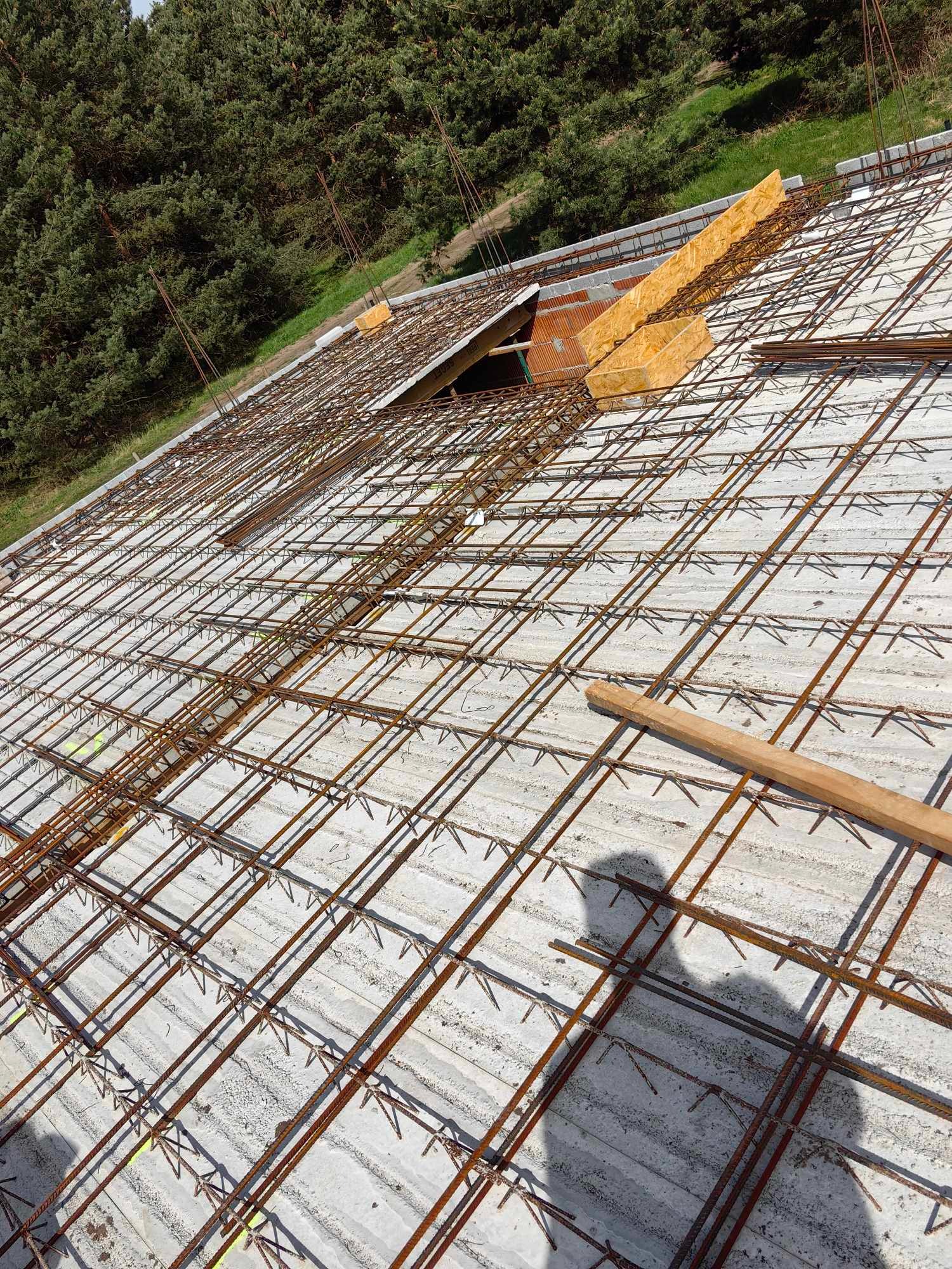 Zbrojenie płyty fundamentowej z widocznym deskowaniem, w tle drzewa i cień osoby.