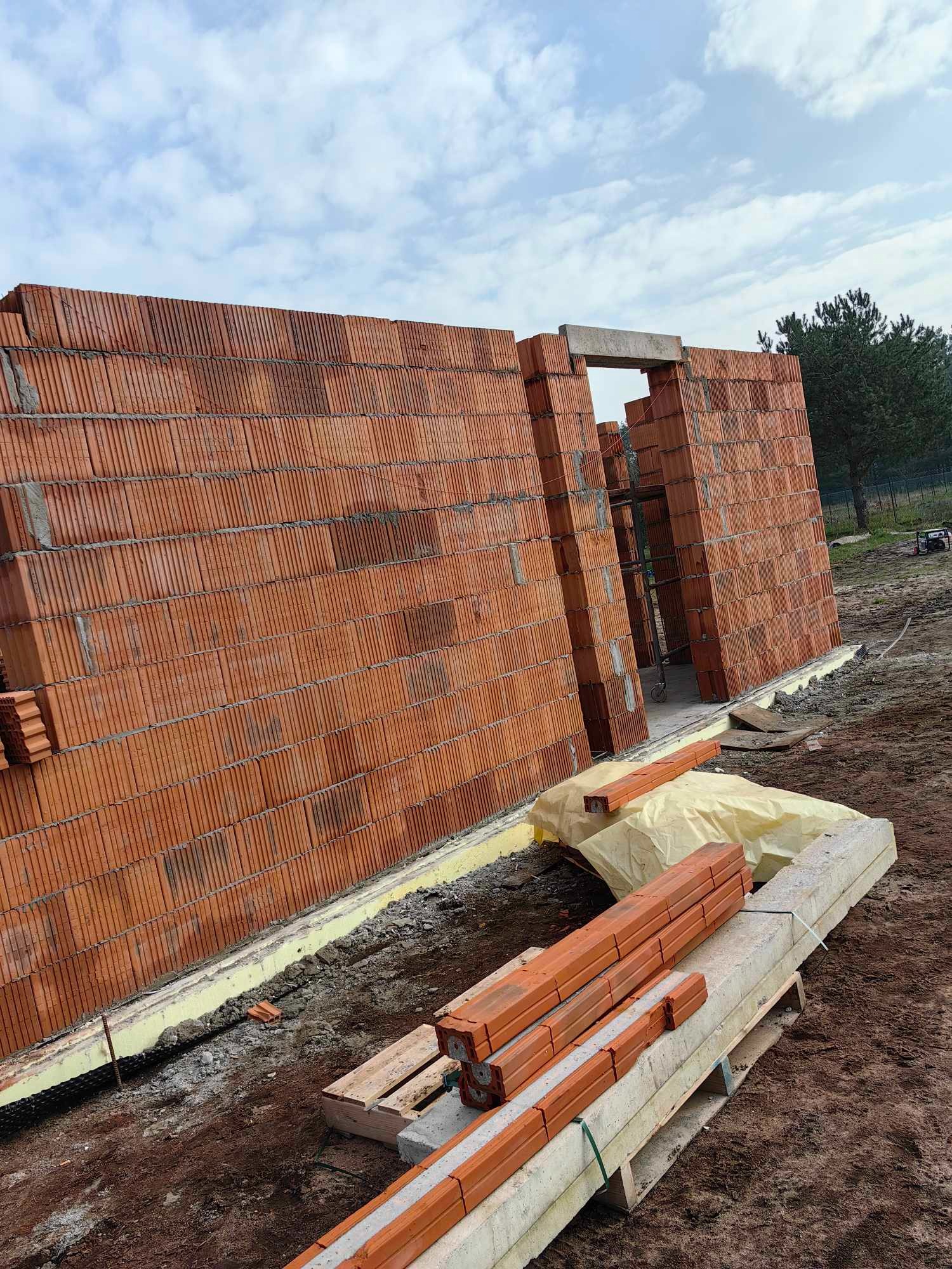 Wznoszenie ścian parterowego domu z czerwonej ceramiki poryzowanej, widoczne nadproże nad otworem drzwiowym, żółta izolacja fundamentów i paleta z bloczkami betonowymi na placu budowy.