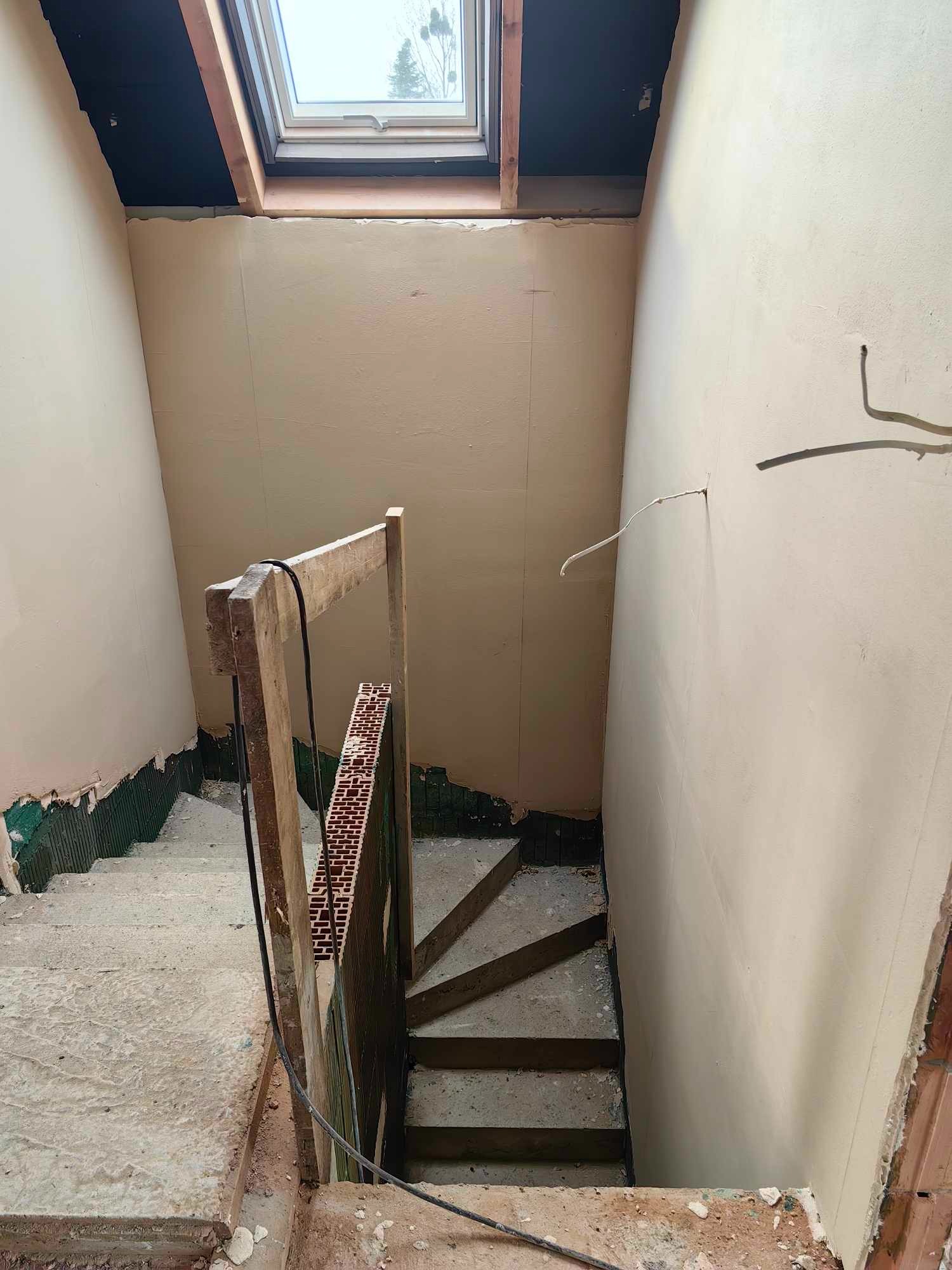 Schody wewnętrzne w trakcie prac wykończeniowych: ściany pokryte świeżo nałożoną gładzią, widoczne drewniane elementy konstrukcyjne schodów i prowizoryczna barierka z cegły, okno dachowe w górnej...