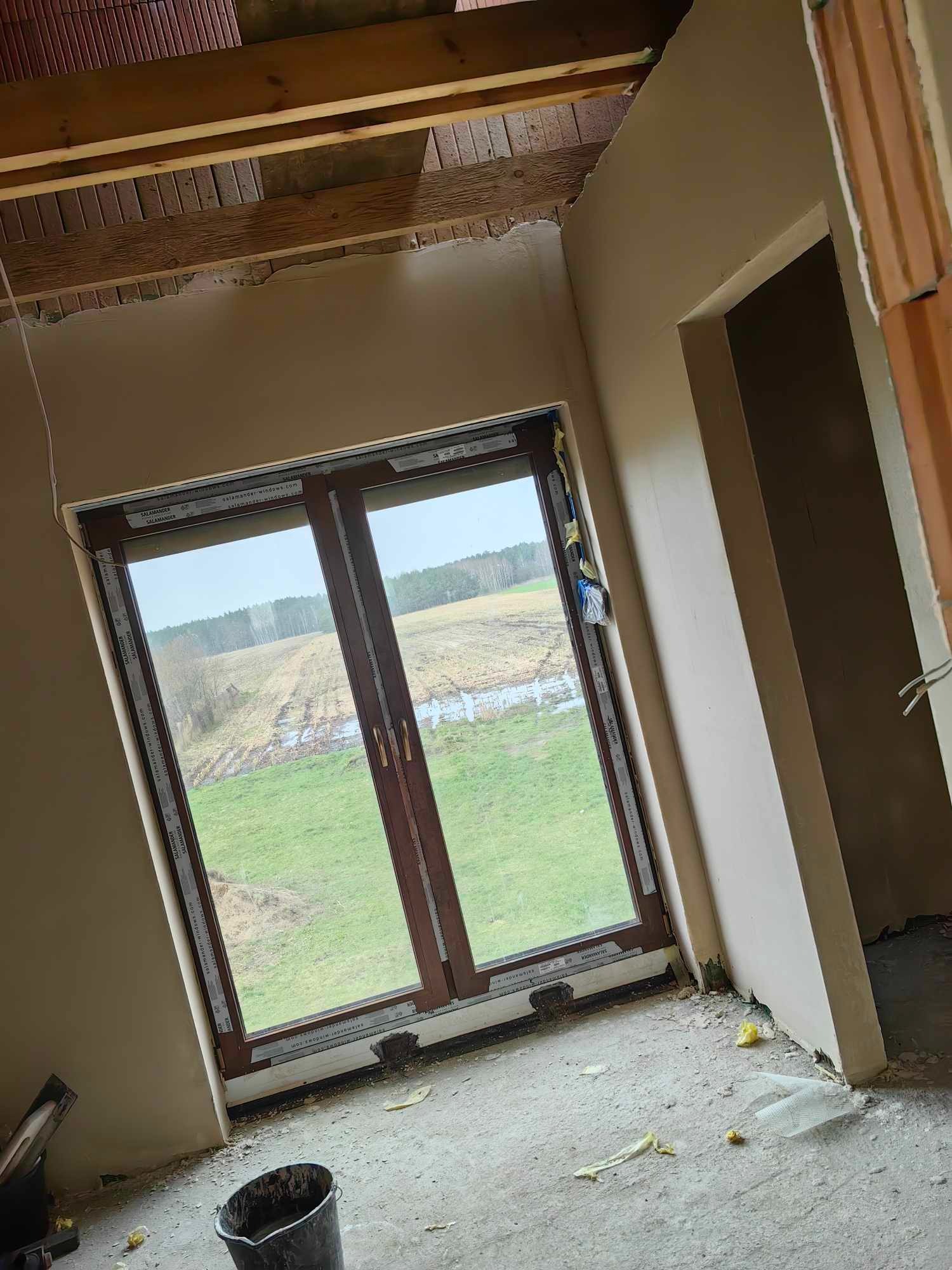 Wnętrze domu w trakcie wykańczania: ściany po nałożeniu gładzi, nowe okno z widokiem na łąkę, surowy strop z drewnianymi belkami, widoczne ślady prac budowlanych.
