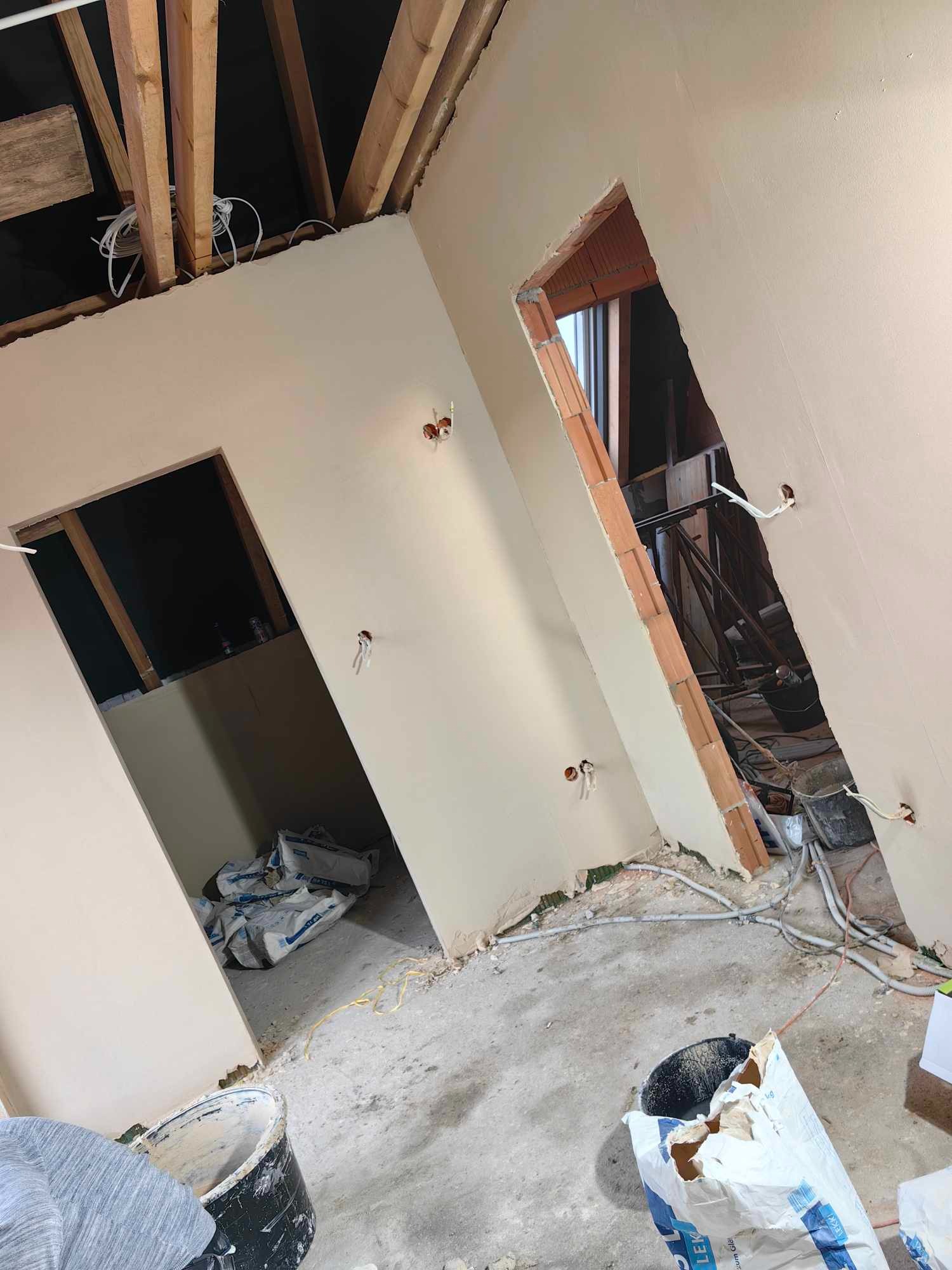 Wnętrze w trakcie prac wykończeniowych: ściany pokryte świeżą warstwą gładzi, widoczne przewody elektryczne wystające ze ścian, surowy strop z drewnianych belek, worki z zaprawą na podłodze...