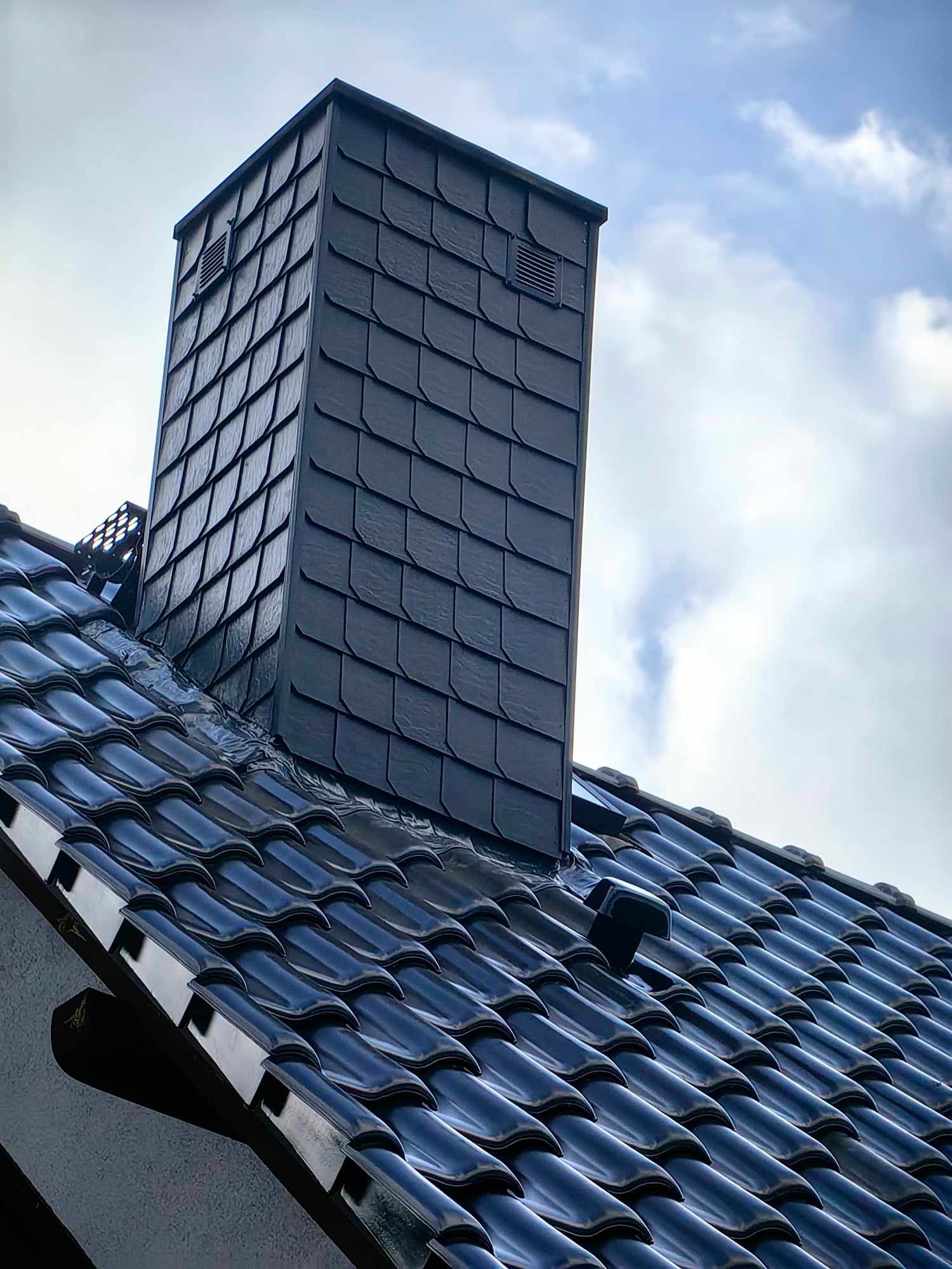 Ciemny, klinkierowy komin z wentylacją, dopasowany kolorystycznie do błyszczącej, ceramicznej dachówki na tle pochmurnego nieba.