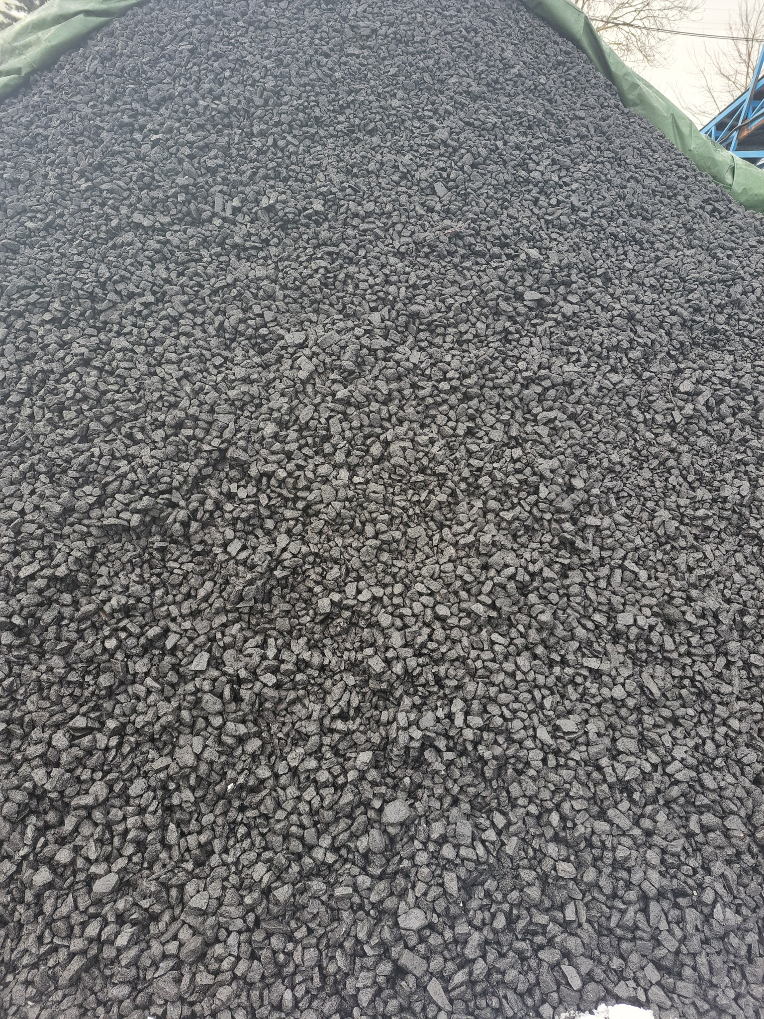 Góra antracytu o drobnej granulacji, częściowo przykryta zieloną plandeką, z widocznymi śladami wilgoci i minimalnym połyskiem.