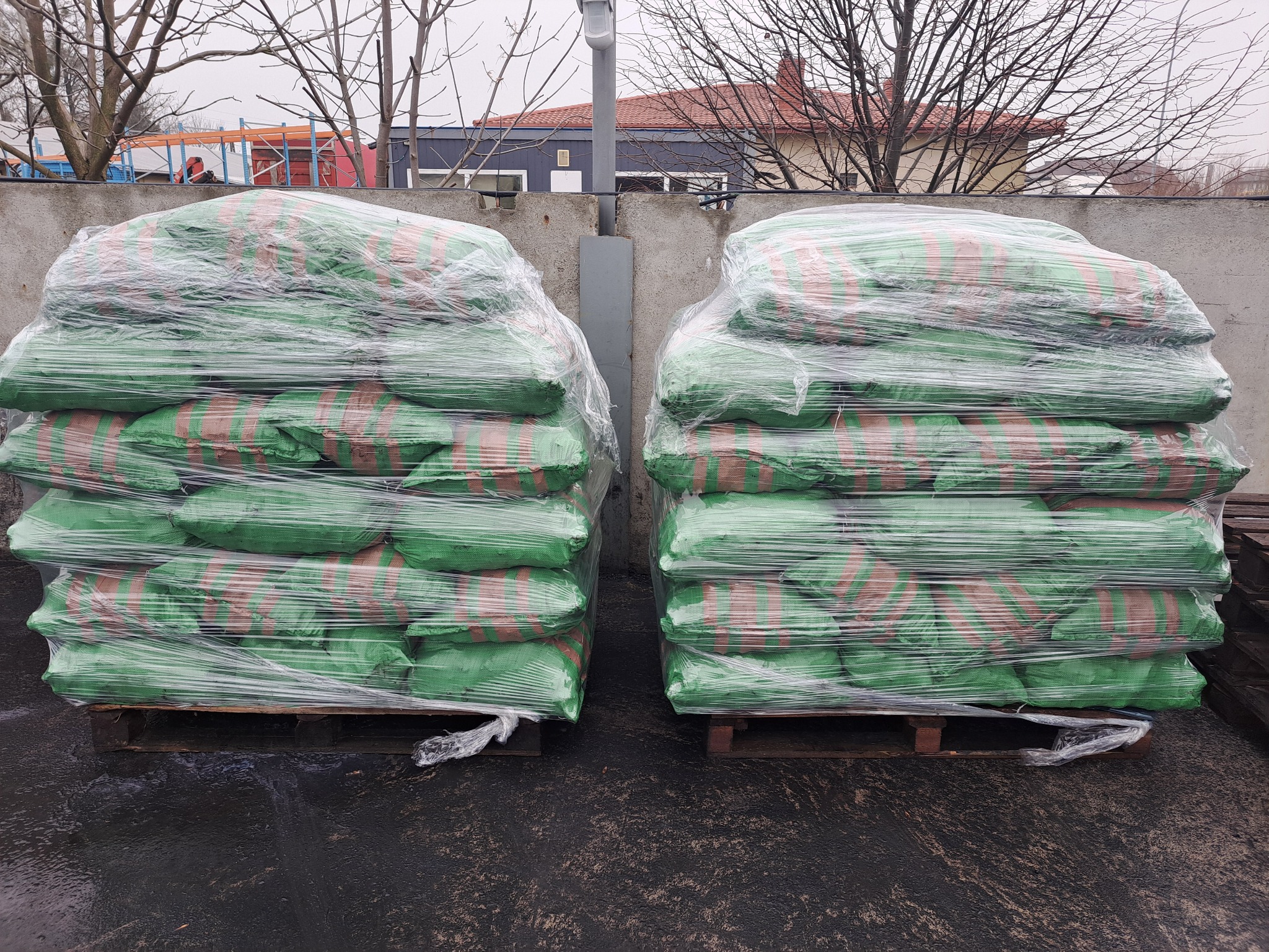 Dwa stosy zielonych worków z brązowymi pasami, owiniętych folią stretch, ułożone na drewnianych paletach na zewnątrz, na tle betonowego muru i budynku z czerwoną dachówką, w pochmurny dzień.