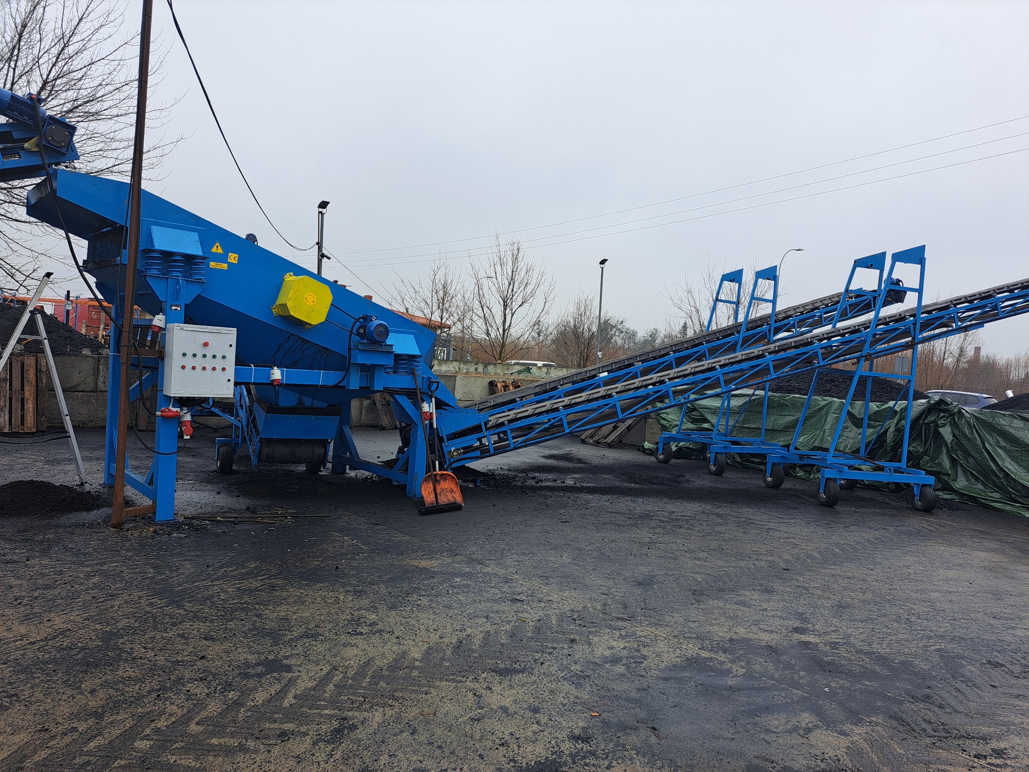Niebieski przenośnik taśmowy do materiałów sypkich na placu, z widoczną pomarańczową łopatą i stertą węgla w tle.