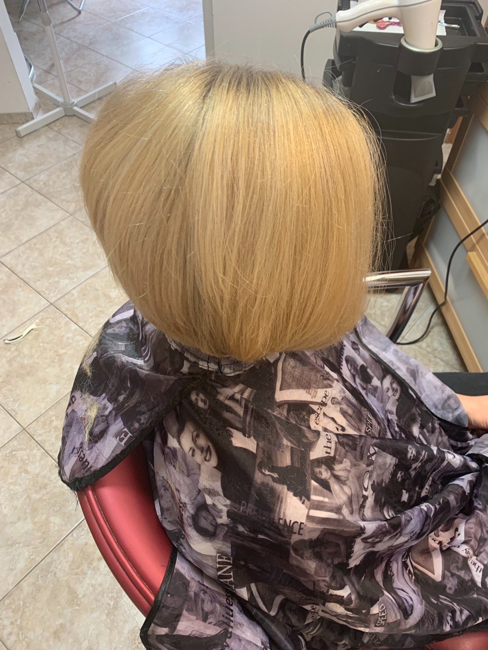 Blond włosy po strzyżeniu bob, widoczne z tyłu, z peleryną ochronną z nadrukiem w stylu retro.