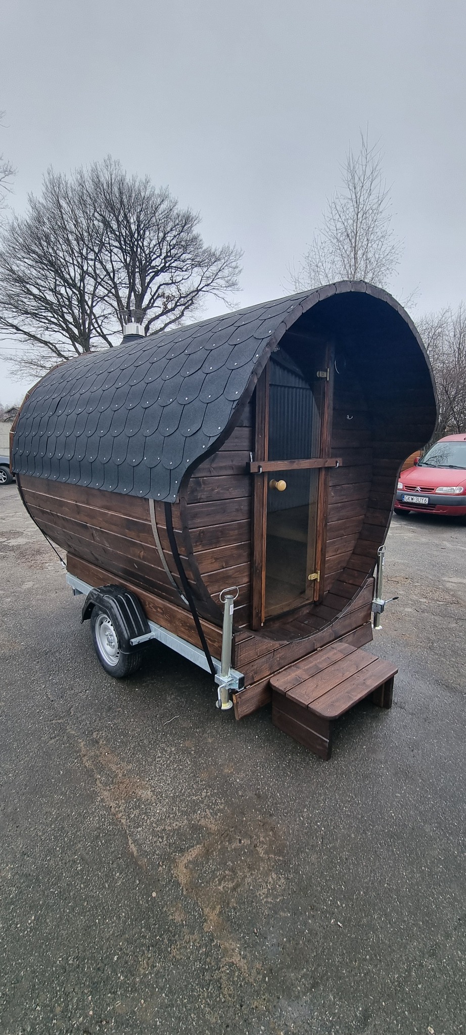 Mobilna sauna w kształcie beczki, z drewnianymi schodkami i dachem z gontu bitumicznego, umieszczona na przyczepie samochodowej. Widoczne drzewa w tle.