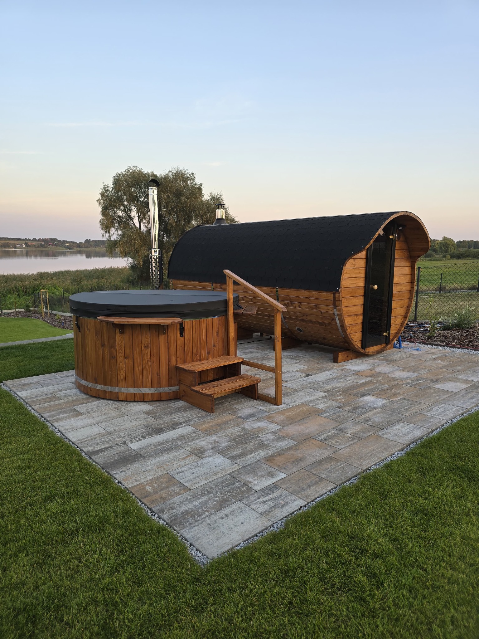 Drewniana sauna beczka i jacuzzi na zewnątrz, na tle jeziora o zmierzchu. Schodki prowadzą do jacuzzi. Całość na wyłożonym kostką placu, otoczona trawnikiem.