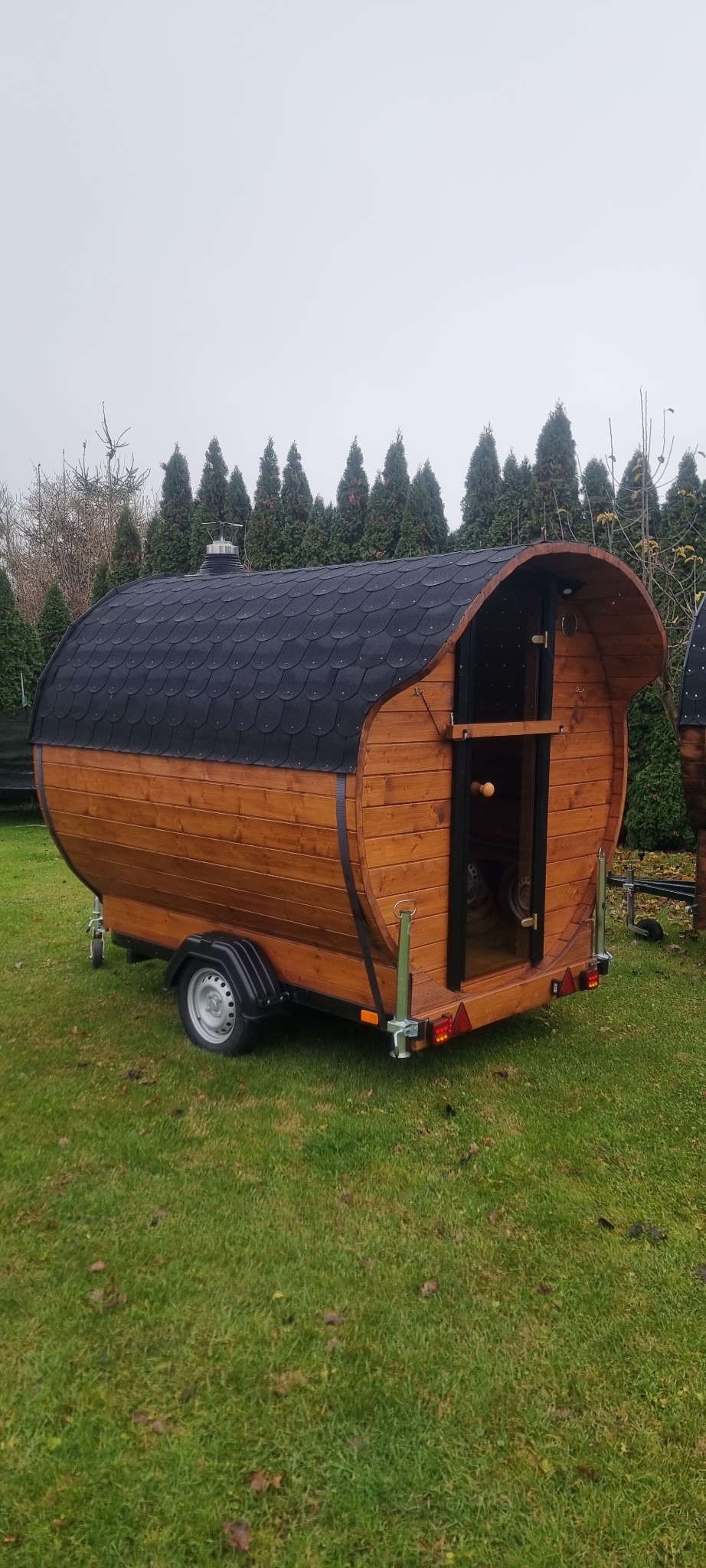Mobilna sauna w kształcie beczki, z drewnianą konstrukcją i czarnym dachem, umieszczona na zielonym trawniku. Widoczne koła i elementy przyczepy.