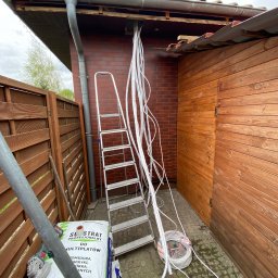 Ruslan Malinin - Montaż przewodów wentylacyjnych na zewnątrz budynku. Widoczna drabina, narzędzia i materiały instalacyjne. Prace w toku, instalacja w trakcie montażu.