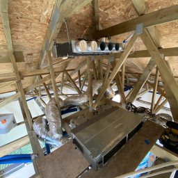 Ruslan Malinin - Montaż rekuperacji w przestrzeni poddasza z widocznymi przewodami wentylacyjnymi, centralą i elementami konstrukcji drewnianej dachu. Ujęcie z dołu.