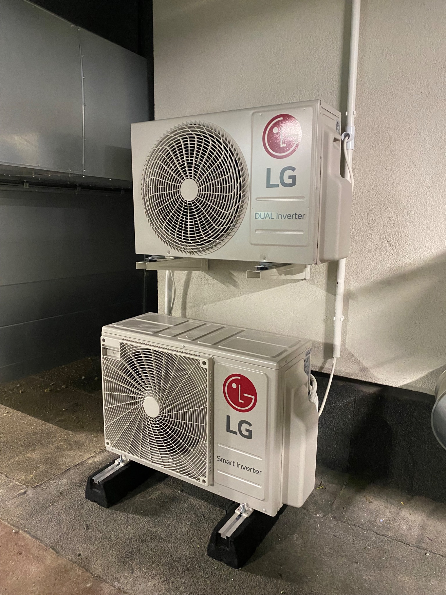 Dwa klimatyzatory LG, jeden nad drugim, zamontowane na zewnątrz budynku. Widoczne przewody i elementy montażowe. Dolny klimatyzator stoi na czarnych podstawkach.