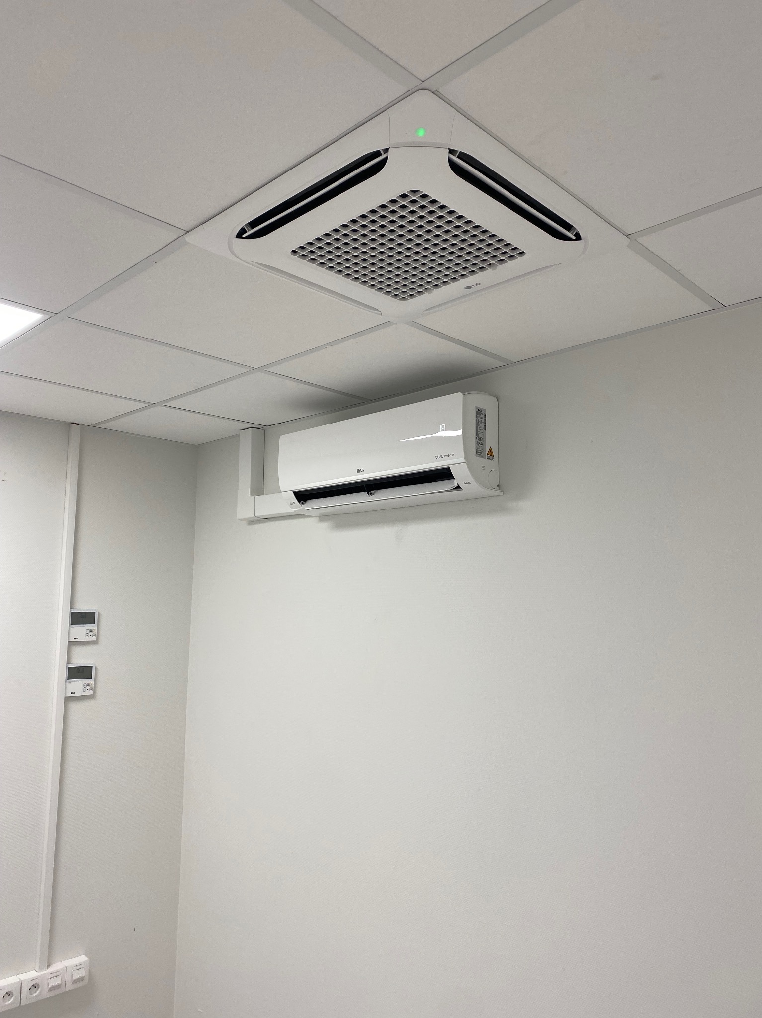 Biała klimatyzacja kasetonowa LG zamontowana w suficie podwieszanym, z widocznym panelem ściennym i jednostką wewnętrzną na ścianie w jasnym pomieszczeniu.