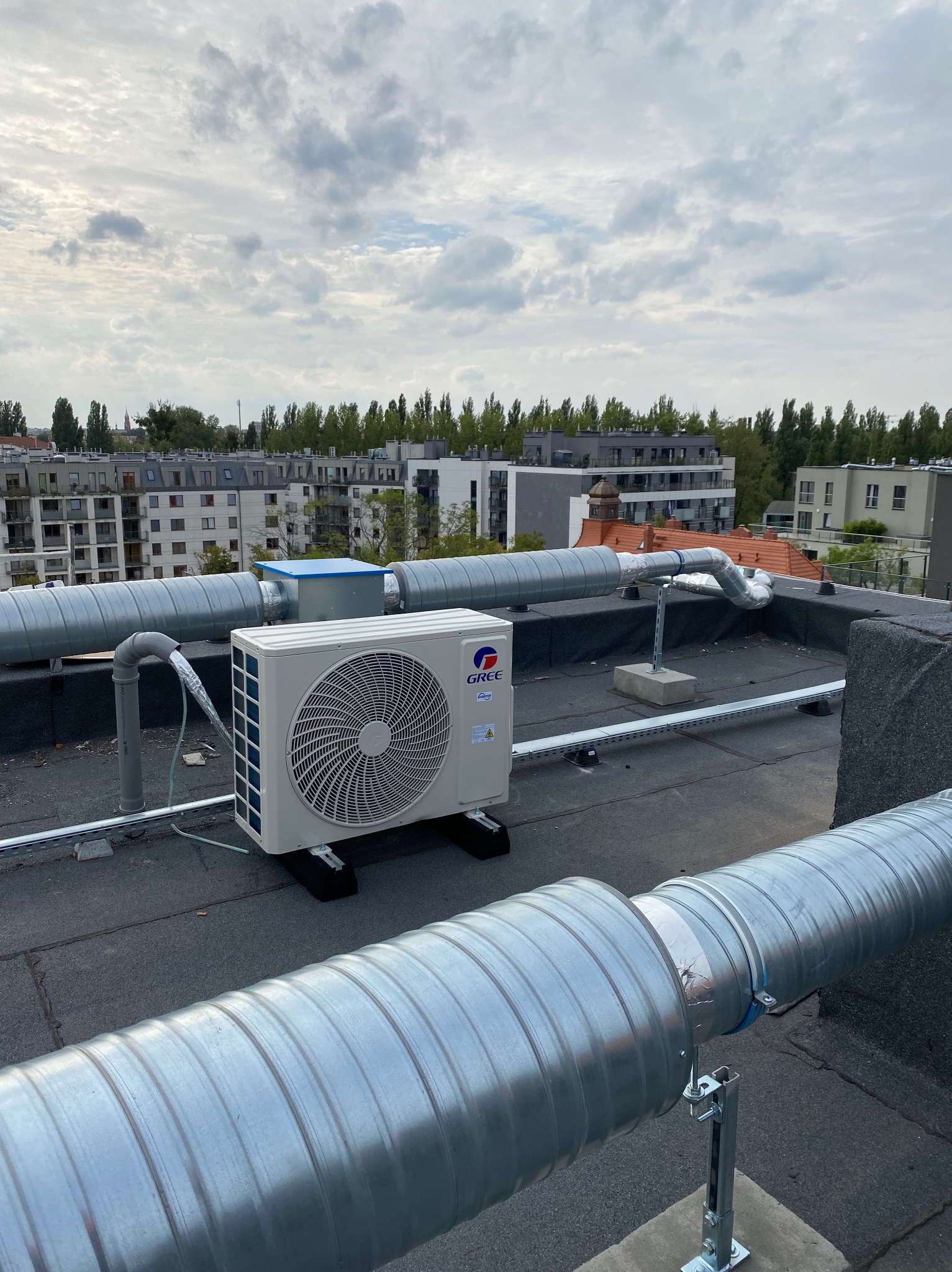 Klimatyzator marki Gree na dachu budynku z widocznymi przewodami wentylacyjnymi. W tle zabudowa miejska i pochmurne niebo.