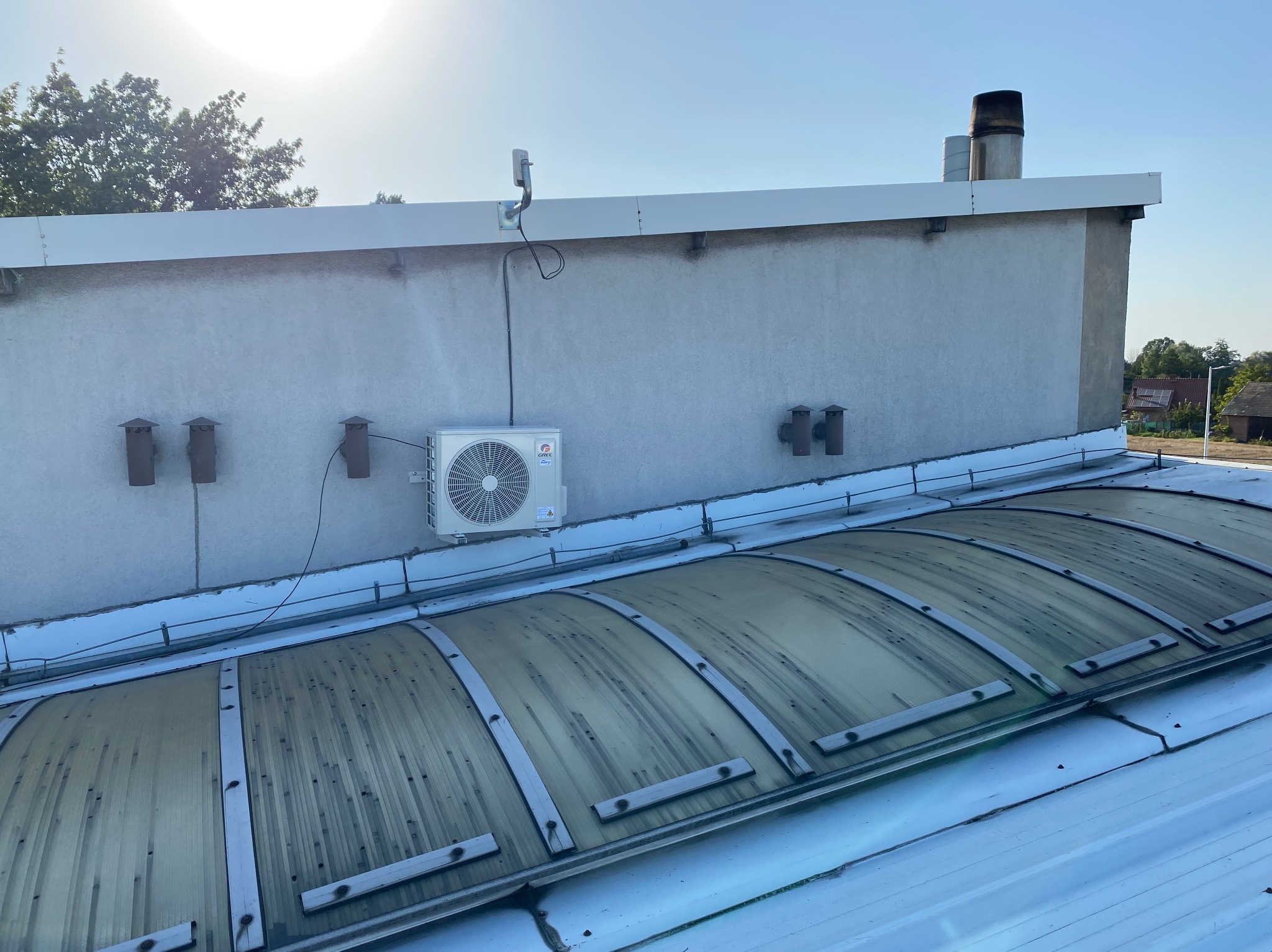 Biała jednostka klimatyzacji GREE zamontowana na szarej ścianie budynku z widocznymi przewodami, anteną i elementami wentylacji. W tle komin i świetlik dachowy.