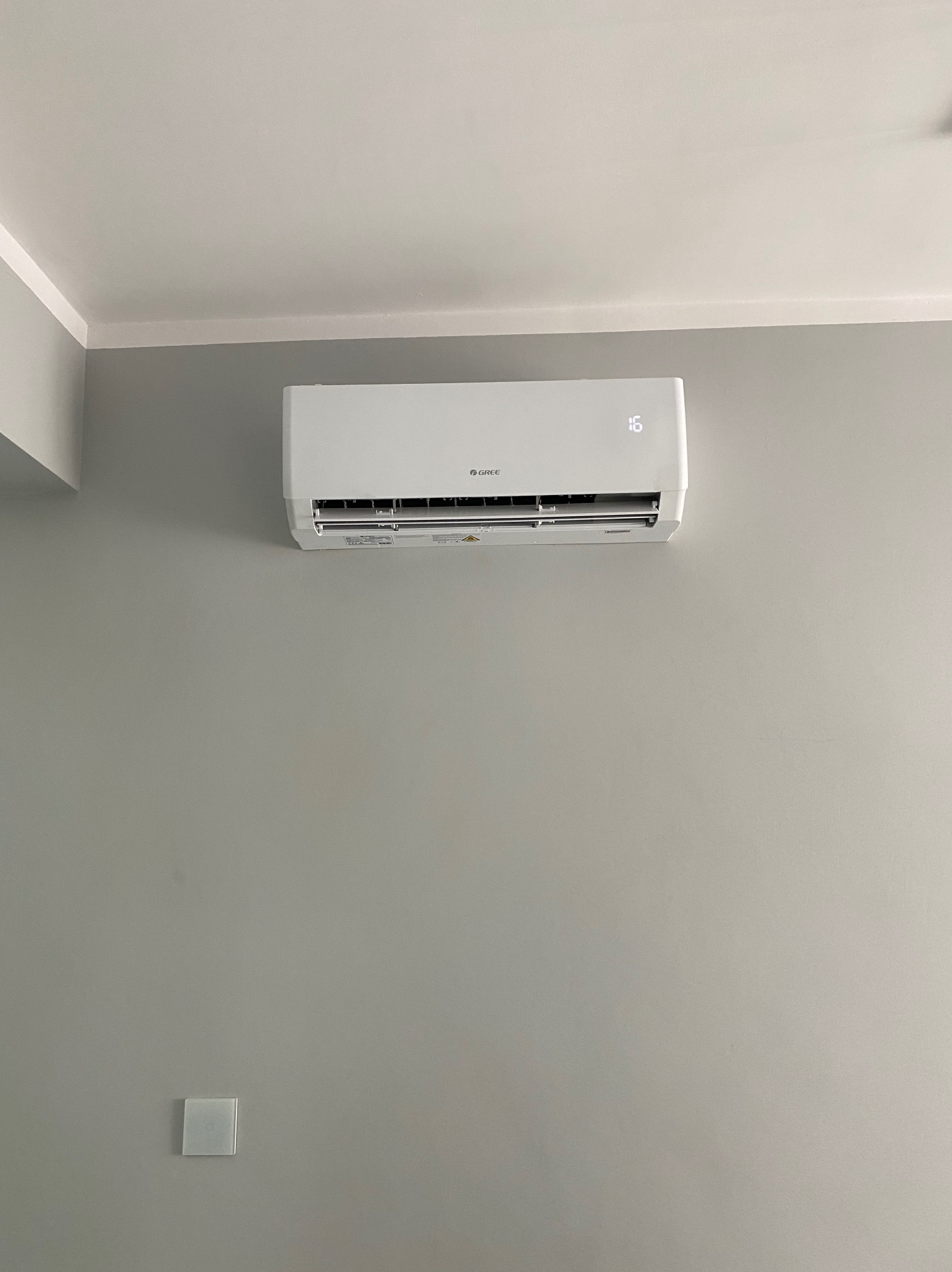 Biała klimatyzacja Gree zamontowana na szarej ścianie w pomieszczeniu. Widoczny wyświetlacz temperatury i panel sterowania. Minimalistyczny design.