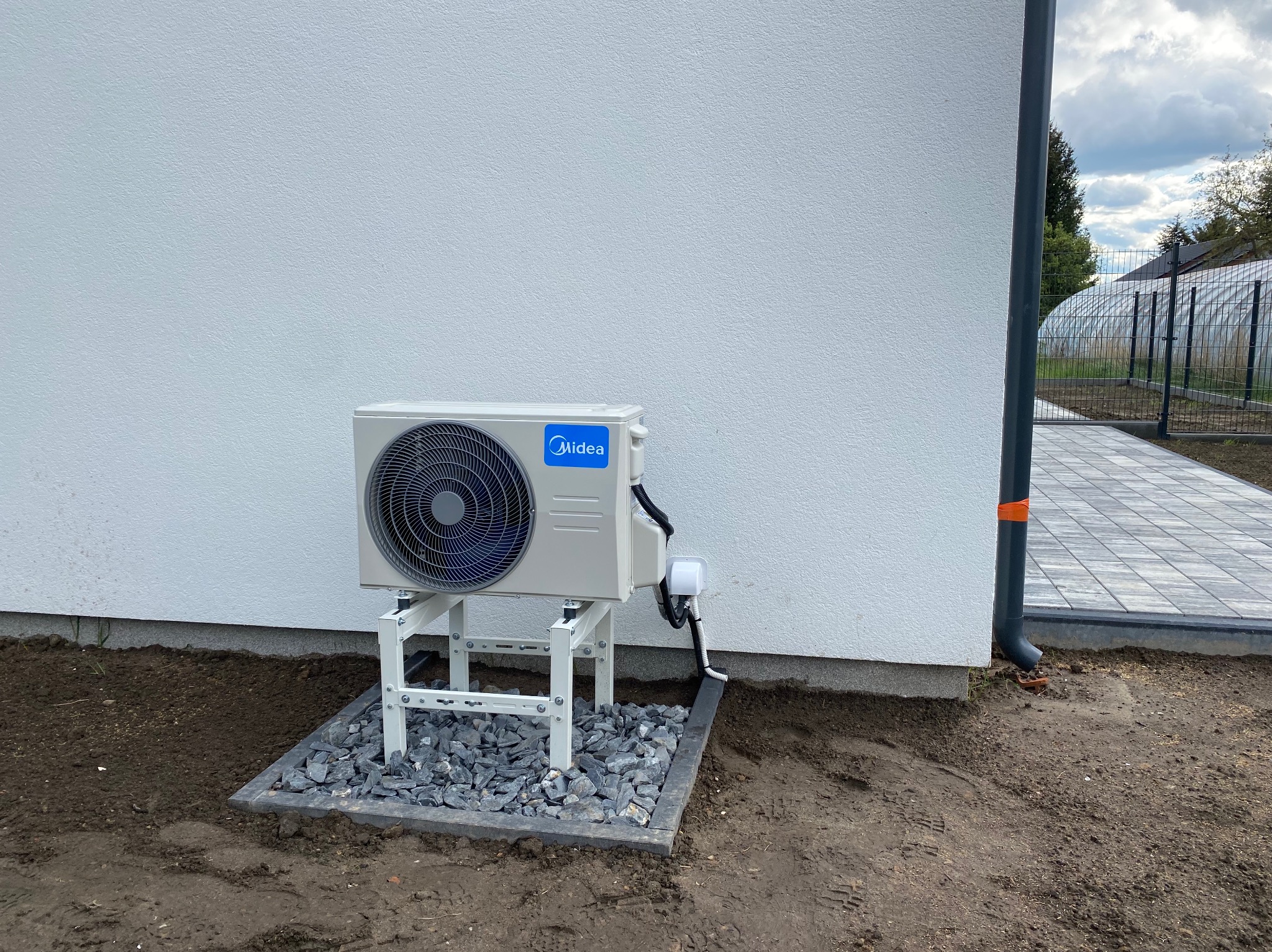 Zewnętrzna jednostka klimatyzacji Midea, zamontowana na metalowym stelażu przy białej ścianie budynku, otoczona kamieniami. Widoczny fragment chodnika i rynna.