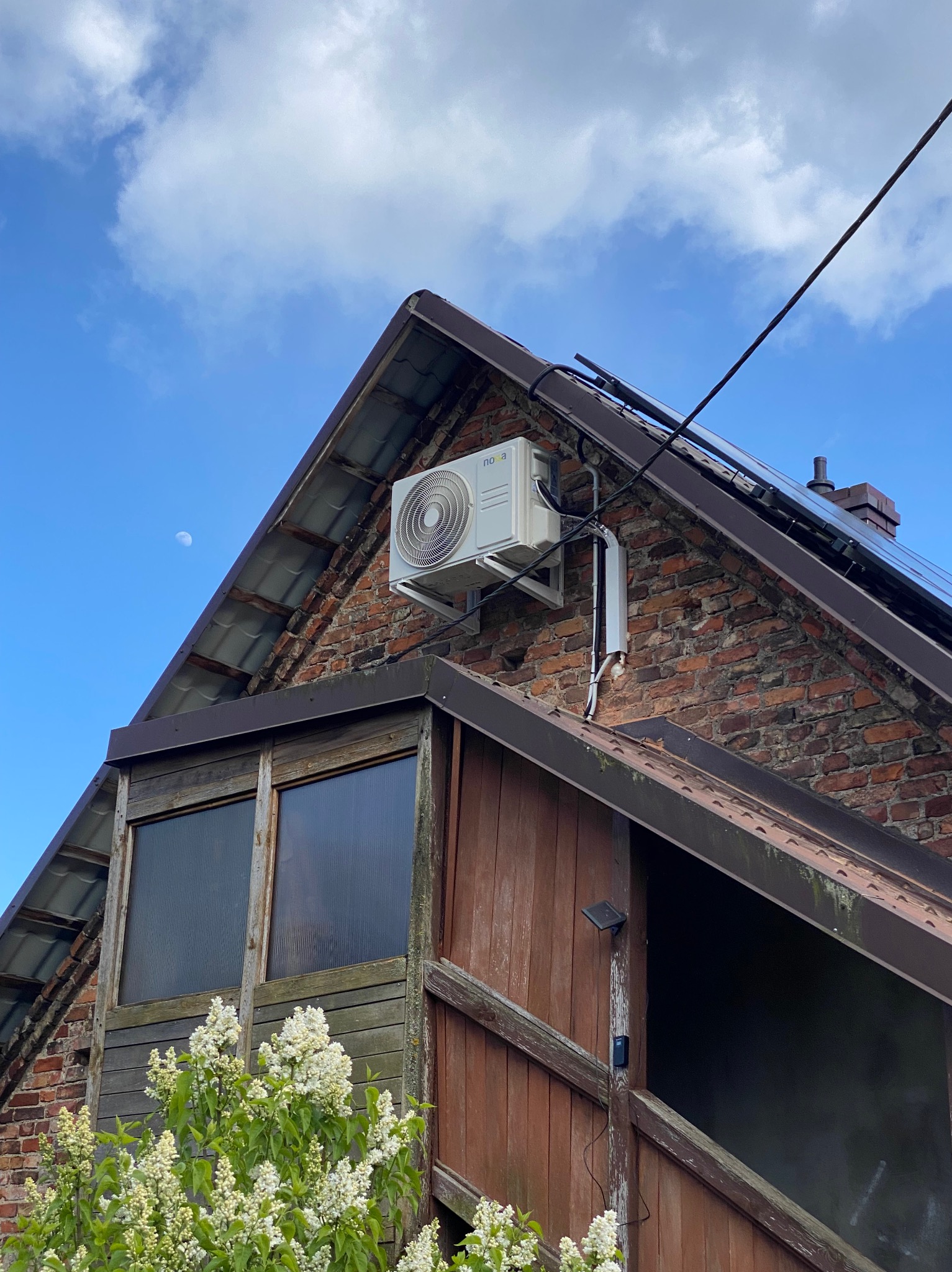 Biała jednostka zewnętrzna klimatyzacji zamontowana na ceglanym szczycie dachu domu, widok z dołu na tle błękitnego nieba z delikatnym księżycem.