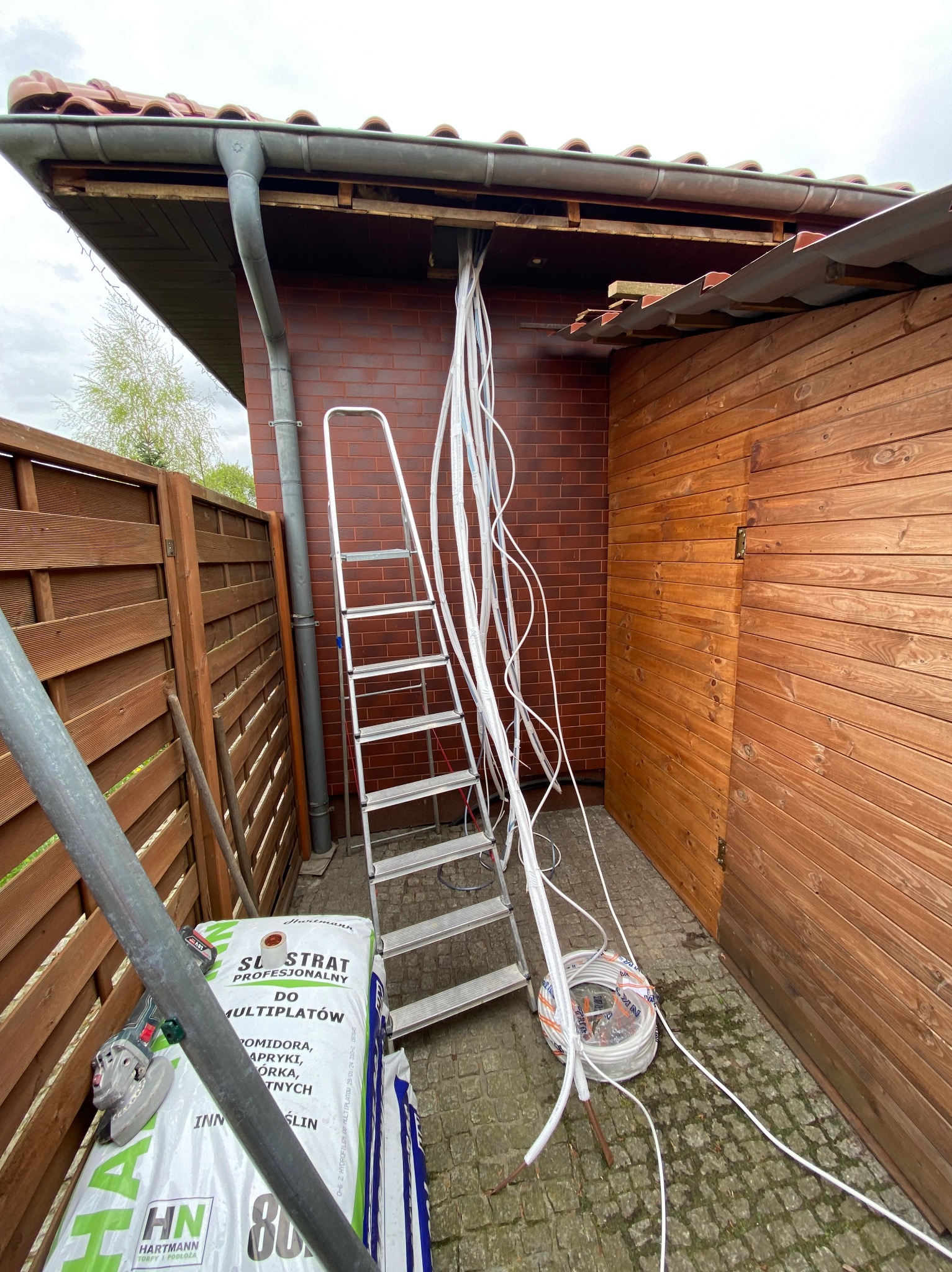 Montaż przewodów wentylacyjnych na zewnątrz budynku. Widoczna drabina, narzędzia i materiały instalacyjne. Prace w toku, instalacja w trakcie montażu.