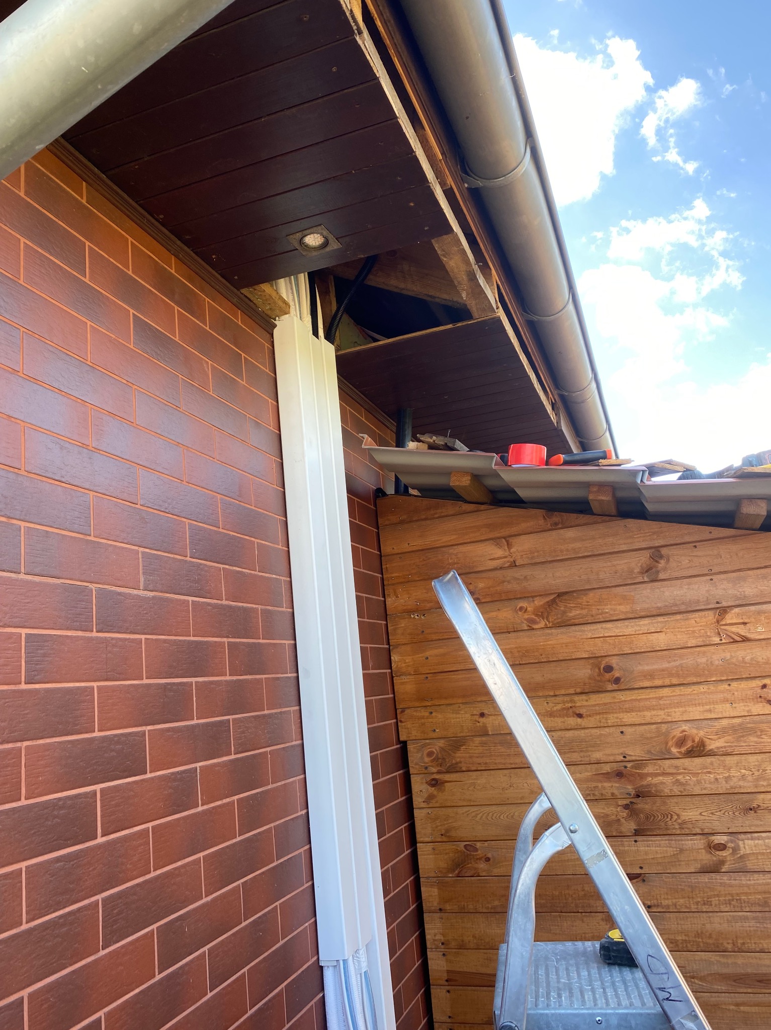 Montaż klimatyzacji zewnętrznej na ceglanej ścianie domu. Widoczna biała rura instalacyjna, drabina i fragment dachu z rynną. Częściowo odsłonięta konstrukcja dachu.