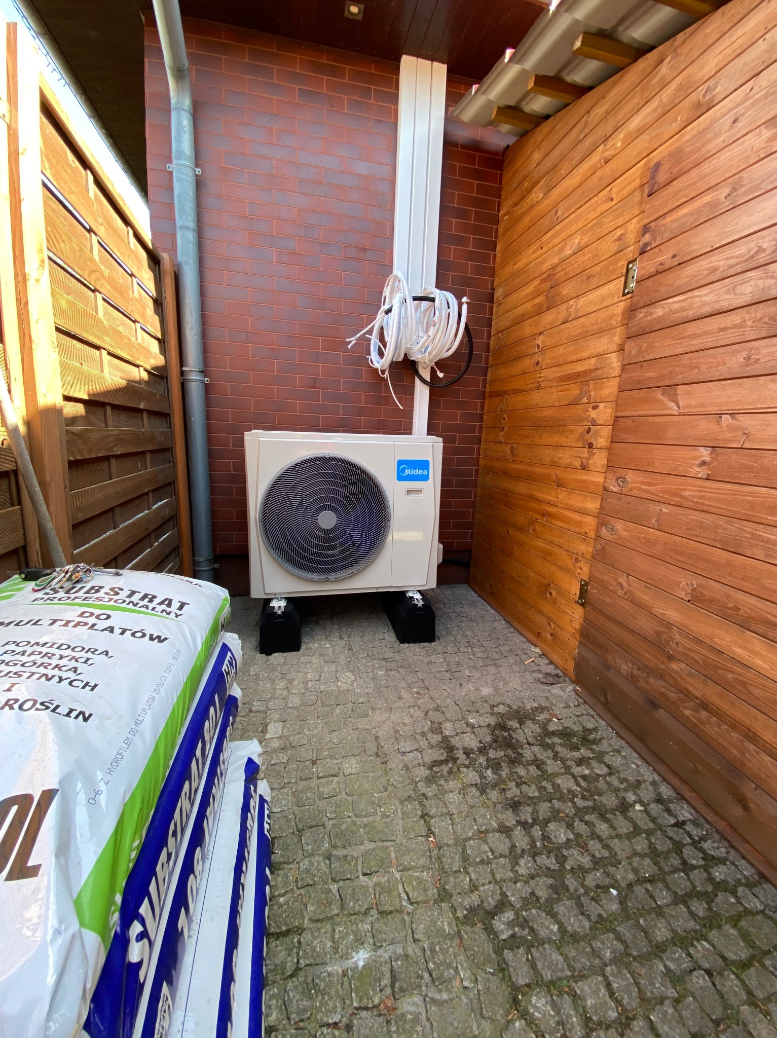 Biała jednostka zewnętrzna klimatyzacji Midea, zamontowana na czarnych podstawach, z przewodami. Obok worki z podłożem do roślin i drewniana ściana.