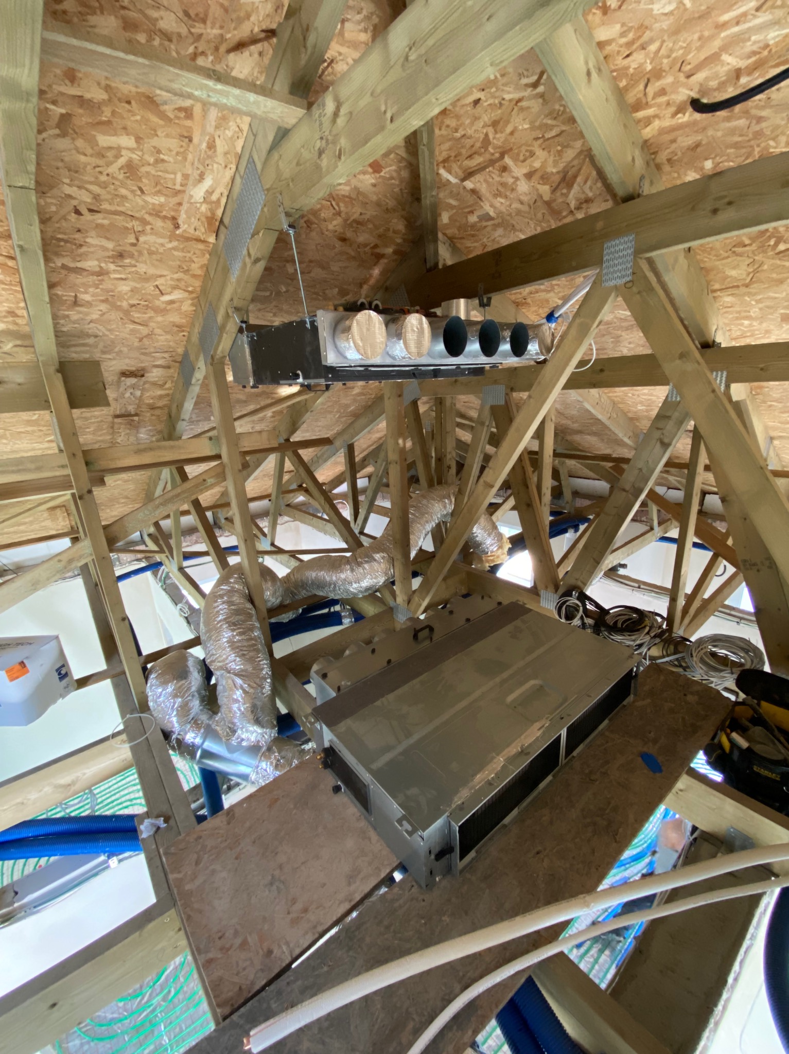 Montaż rekuperacji w przestrzeni poddasza z widocznymi przewodami wentylacyjnymi, centralą i elementami konstrukcji drewnianej dachu. Ujęcie z dołu.