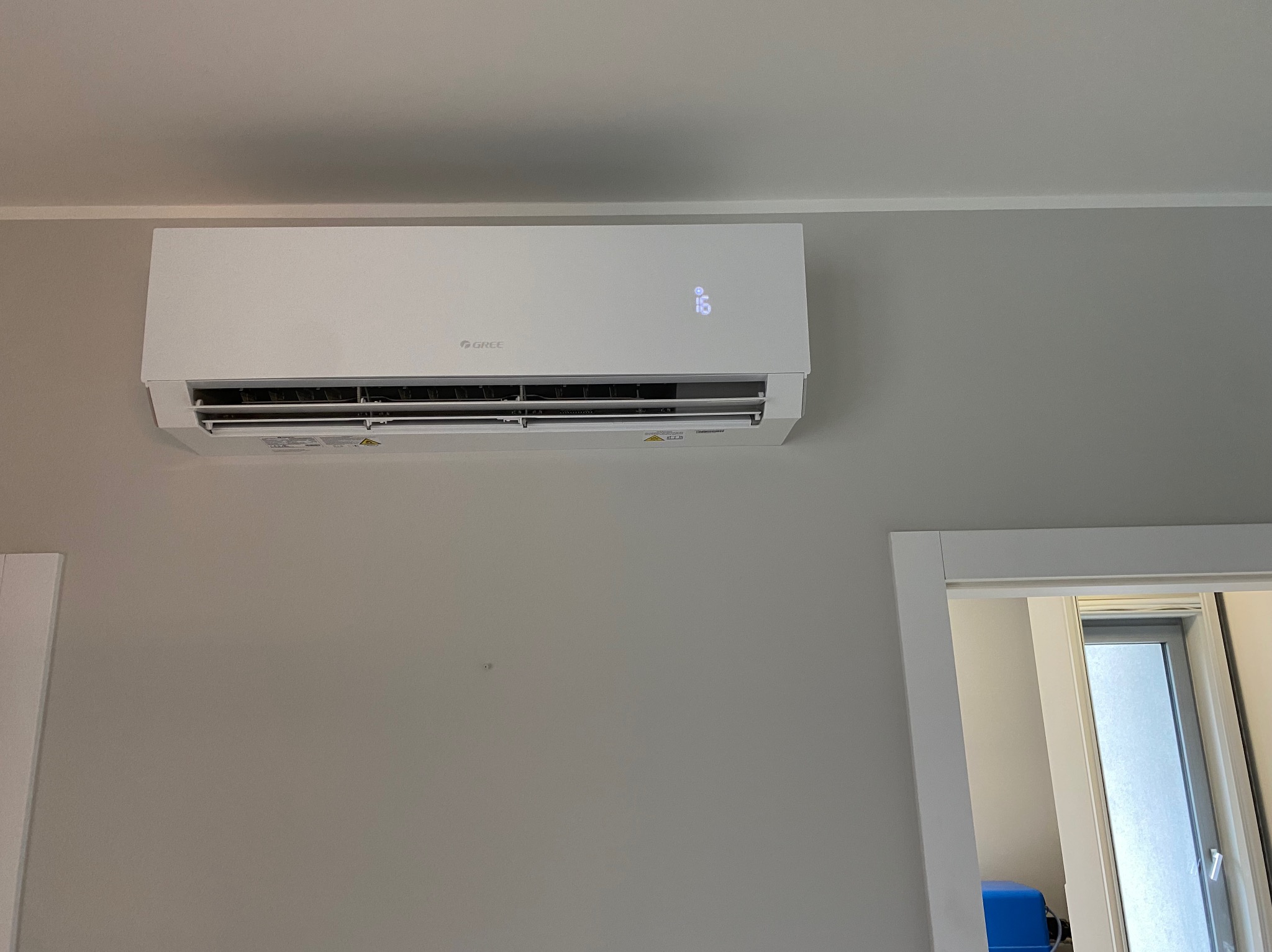 Biała klimatyzacja marki Gree zamontowana na szarej ścianie w pomieszczeniu. Widoczny fragment futryny drzwi. Na klimatyzatorze wyświetla się temperatura 16 stopni.