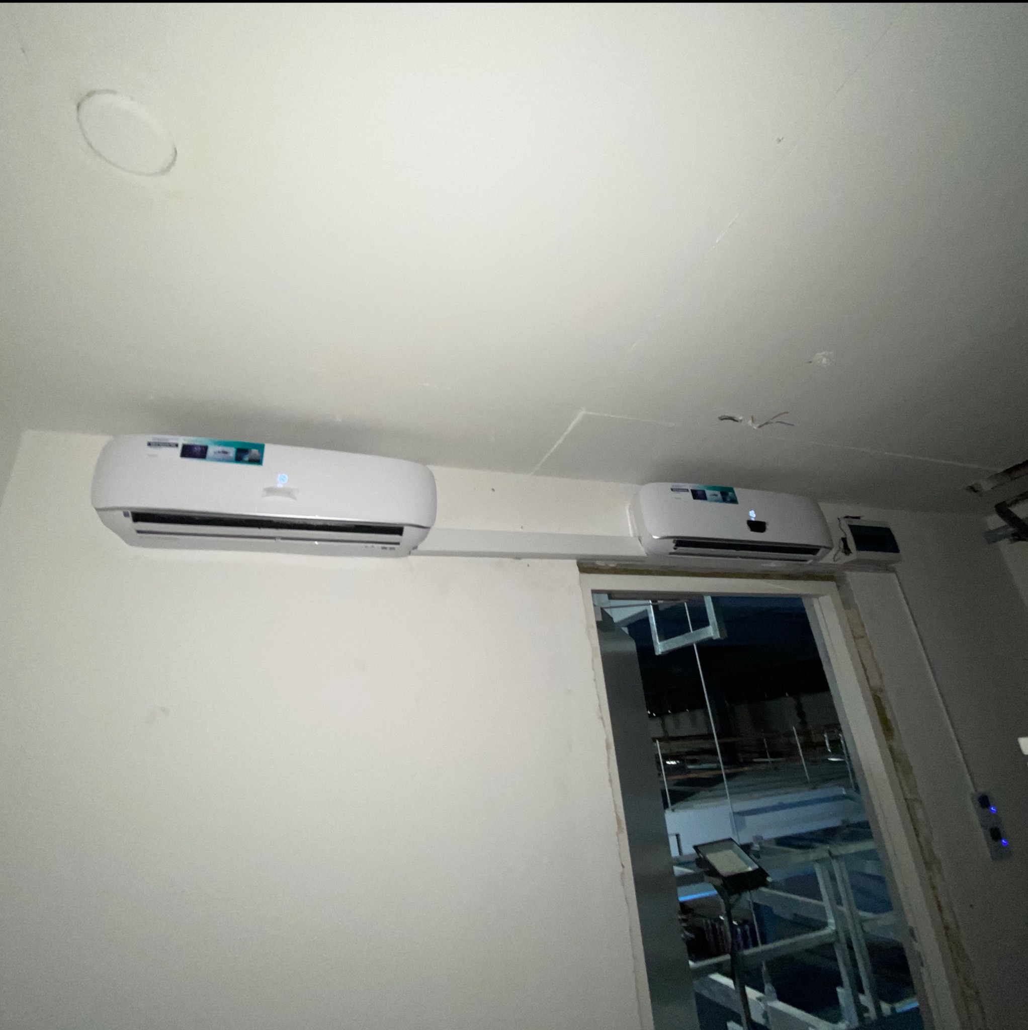 Dwa białe klimatyzatory ścienne zamontowane obok siebie w jasnym pomieszczeniu, widoczne przez otwarte drzwi. Nierówne wykończenie ściany.