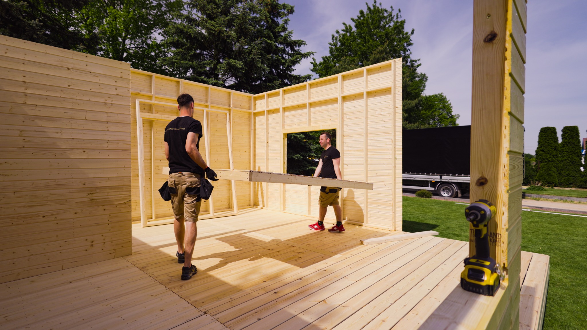 Montaż konstrukcji z drewna: Dwóch mężczyzn przenosi element ściany z oknem wewnątrz częściowo zmontowanej drewnianej konstrukcji z desek, widoczny wkrętak elektryczny, trawa i drzewa w tle.