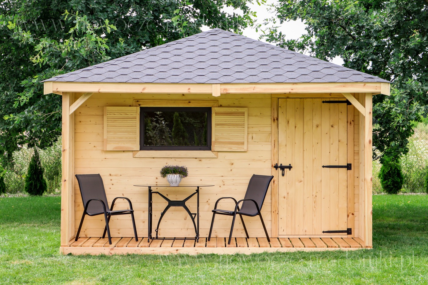 Mała, drewniana konstrukcja z dwuspadowym dachem krytym gontem bitumicznym, z oknem z drewnianymi okiennicami i drzwiami z czarnymi zawiasami, wyposażona w stolik i dwa krzesła na drewnianym...