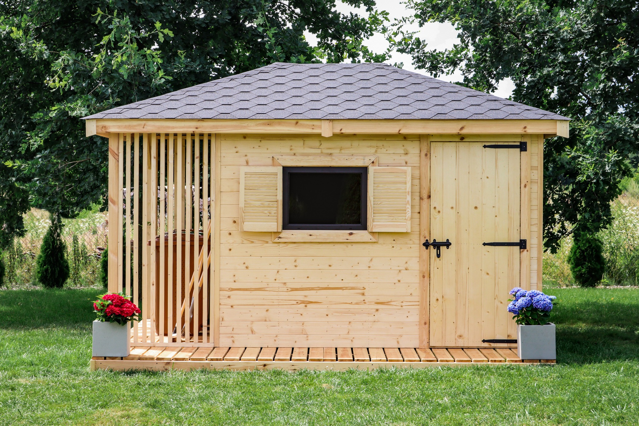 Mała, drewniana konstrukcja z szarym dachem, prawdopodobnie domek narzędziowy lub mała altana, z otwartym wejściem z jednej strony i drzwiami z drugiej, udekorowana kwiatami w donicach po obu...