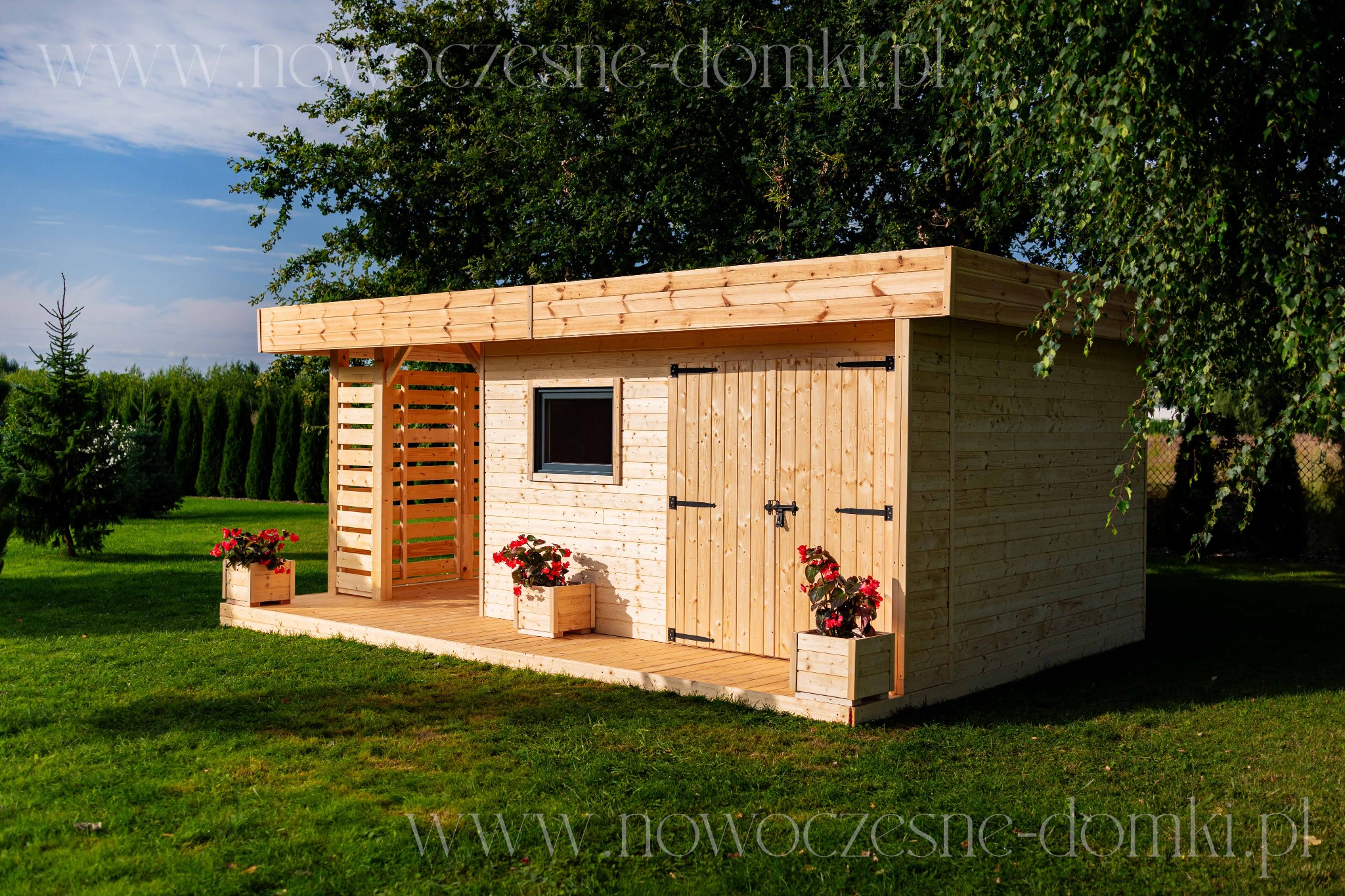 Drewniana konstrukcja z małym oknem i drzwiami na tle zielonego trawnika i drzew, ozdobiona czerwonymi kwiatami w donicach.