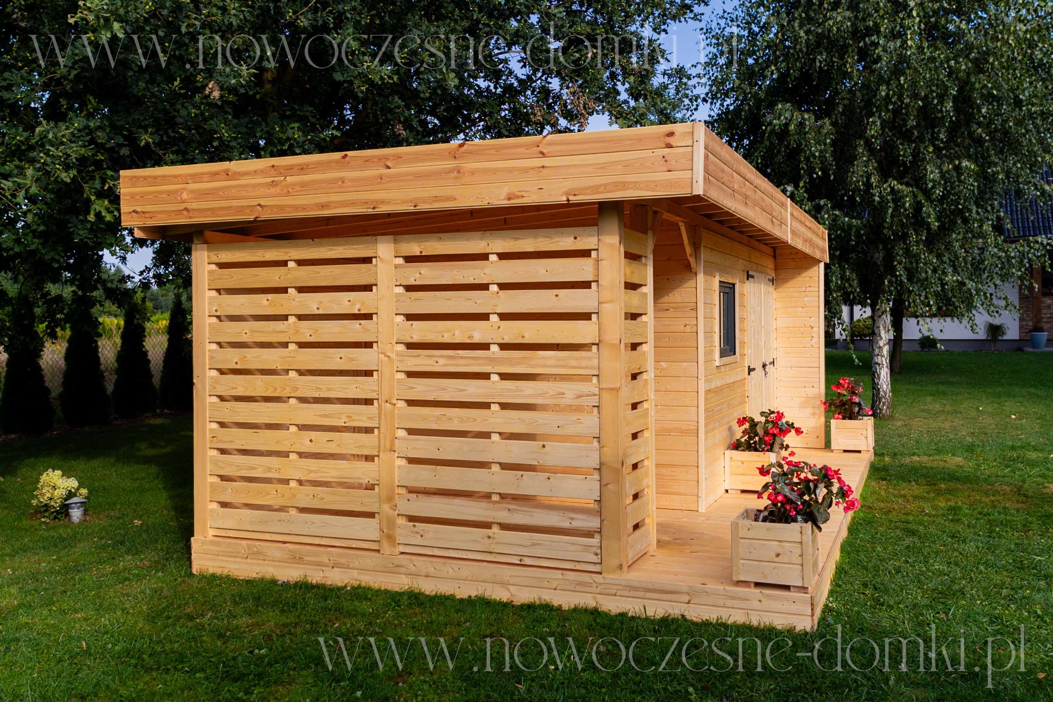 Drewniana konstrukcja z ażurowymi ścianami i płaskim dachem, otoczona zielonym trawnikiem i drzewami, z dwoma donicami z czerwonymi kwiatami na podeście.