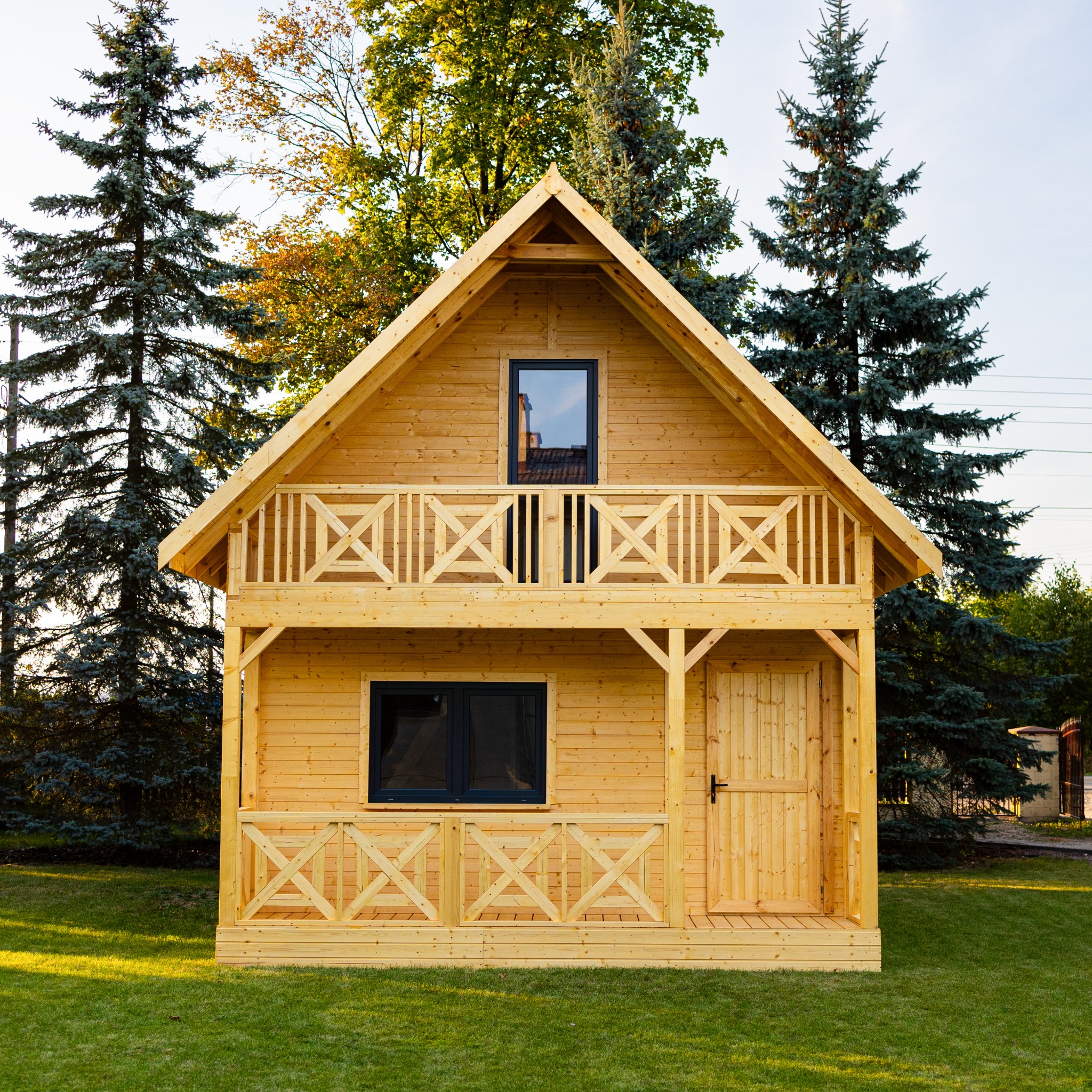 Drewniany domek letniskowy z balkonem w Nieznanicach, wykonany z jasnego drewna, otoczony zielonym trawnikiem i iglastymi drzewami.