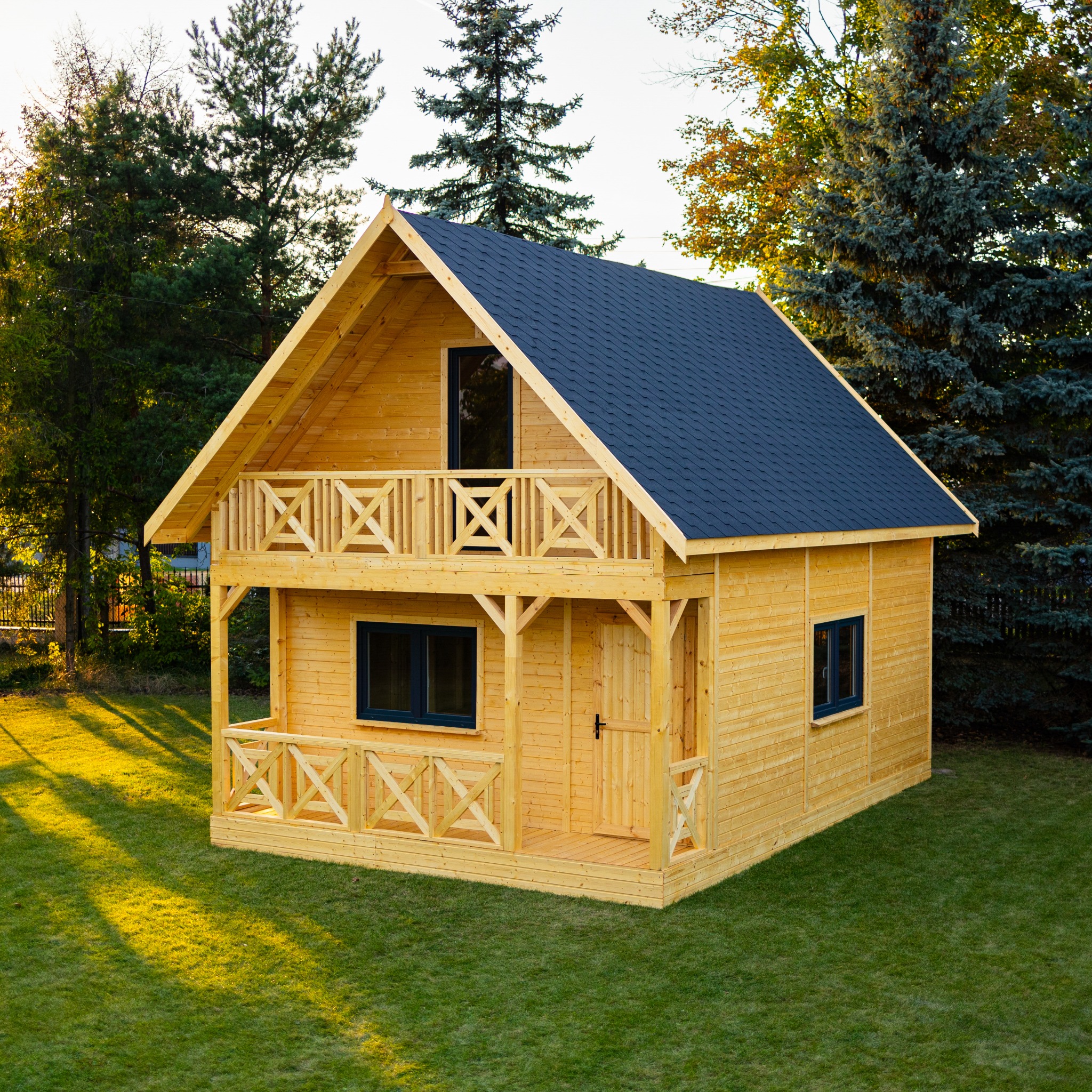 Dwukondygnacyjny domek z drewna o jasnej elewacji, z balkonem i ciemnym dachem, otoczony zielonym trawnikiem i drzewami iglastymi.