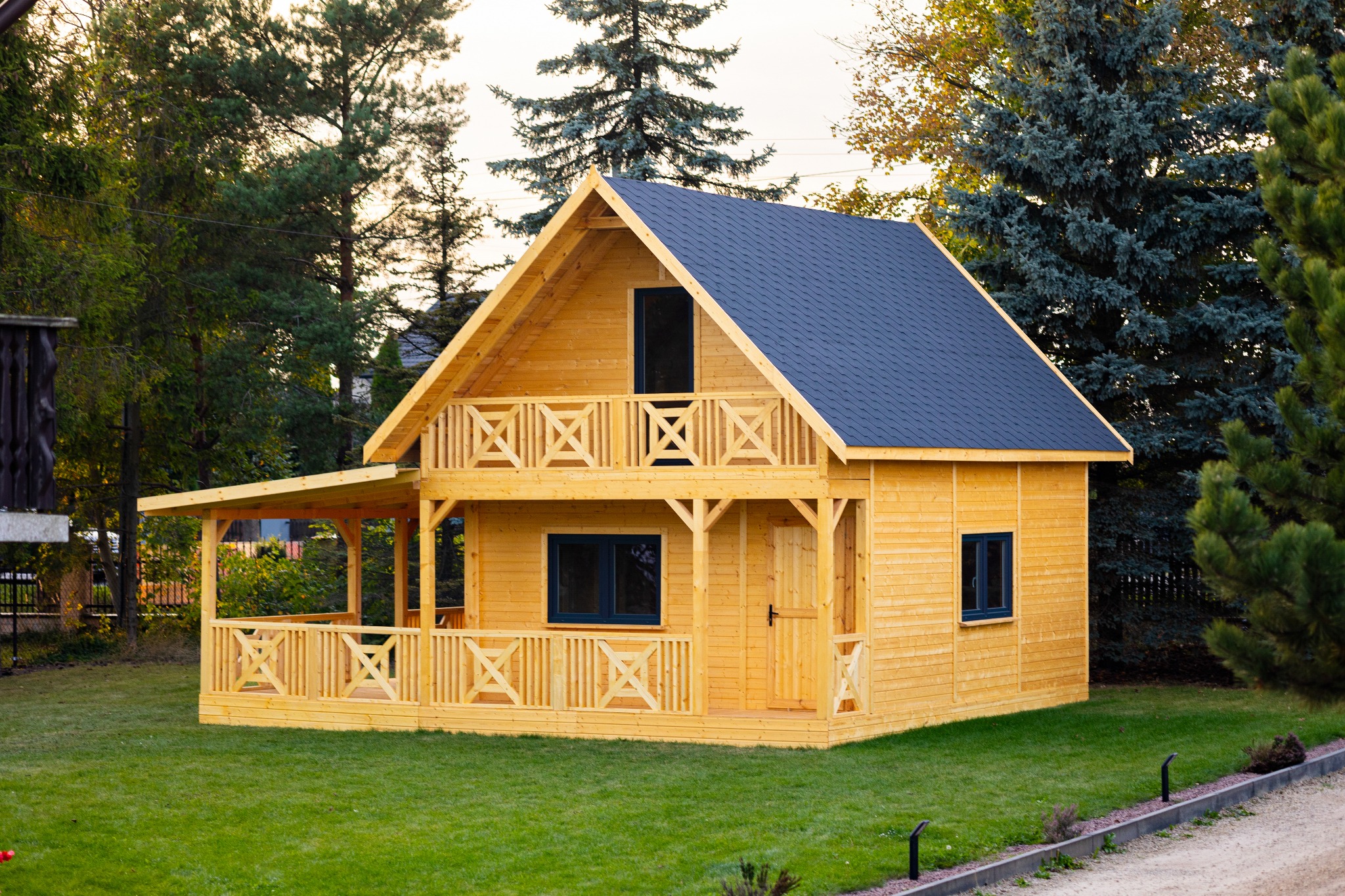 Dwukondygnacyjny, drewniany domek letniskowy z ciemnym dachem, balkonem i zadaszonym tarasem, otoczony zielonym trawnikiem i drzewami.