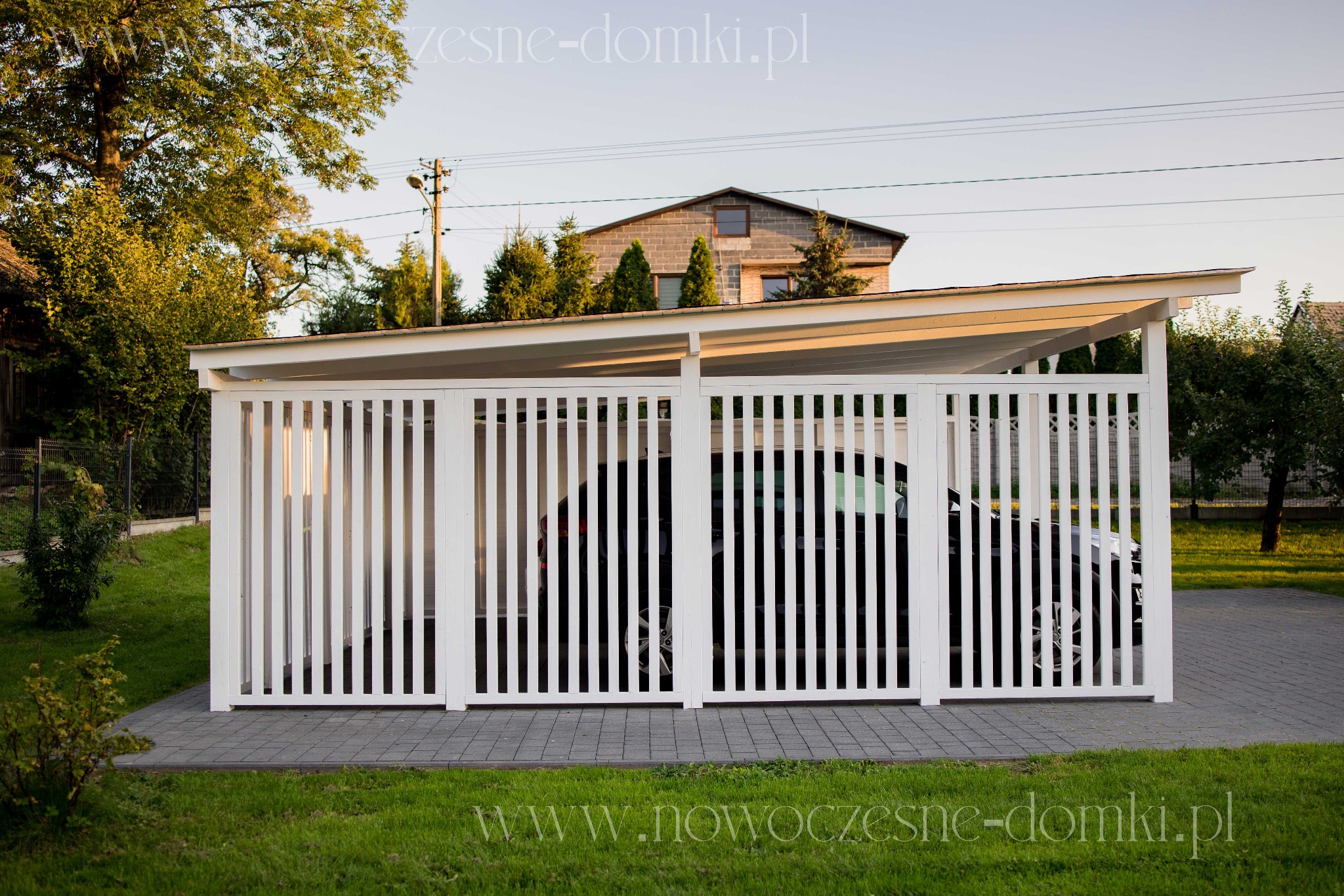 Nowoczesna, biała konstrukcja z pionowych listew, pełniąca funkcję zadaszonego miejsca parkingowego, z widocznym samochodem wewnątrz, na tle domu jednorodzinnego i zieleni.