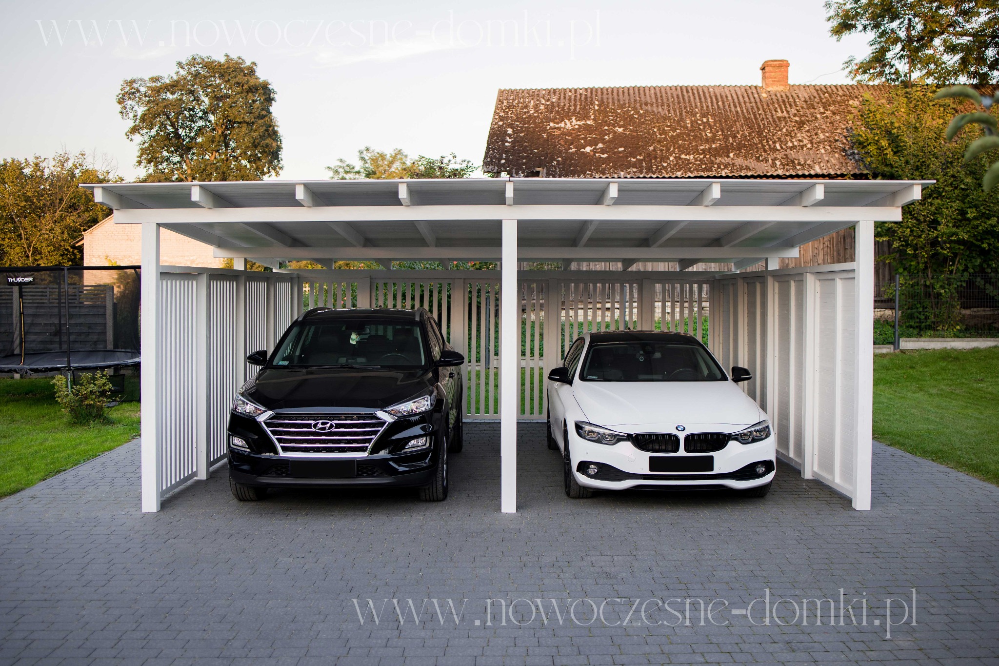 Biała, drewniana wiata garażowa z pionowymi listwami, mieszcząca czarny Hyundai Tucson i białe BMW, na posesji z kostki brukowej.