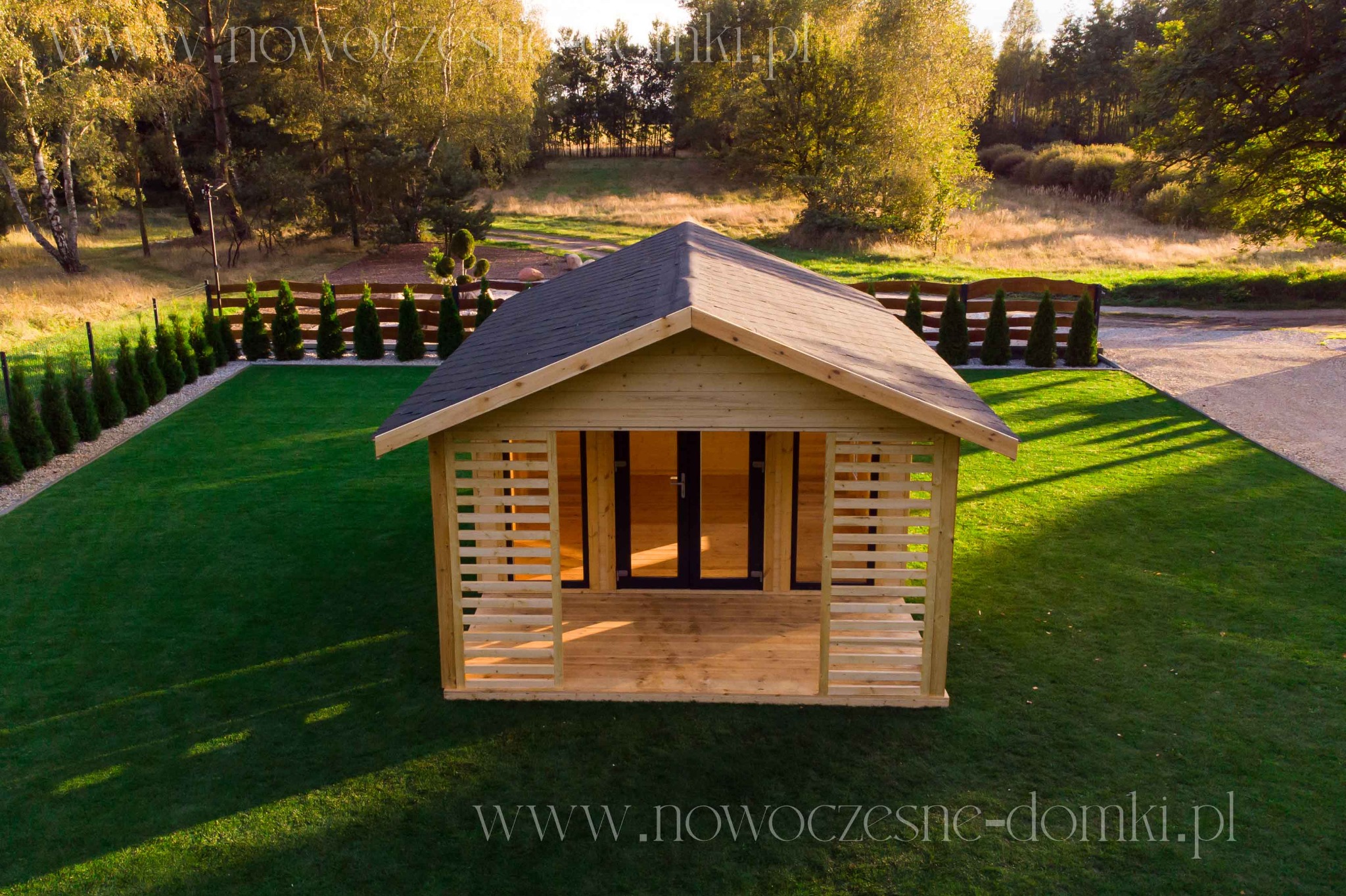 Nowoczesna, drewniana altana z płaskim dachem i przeszklonymi drzwiami, otoczona idealnie przystrzyżonym trawnikiem i żywopłotem z iglaków, widok z góry.