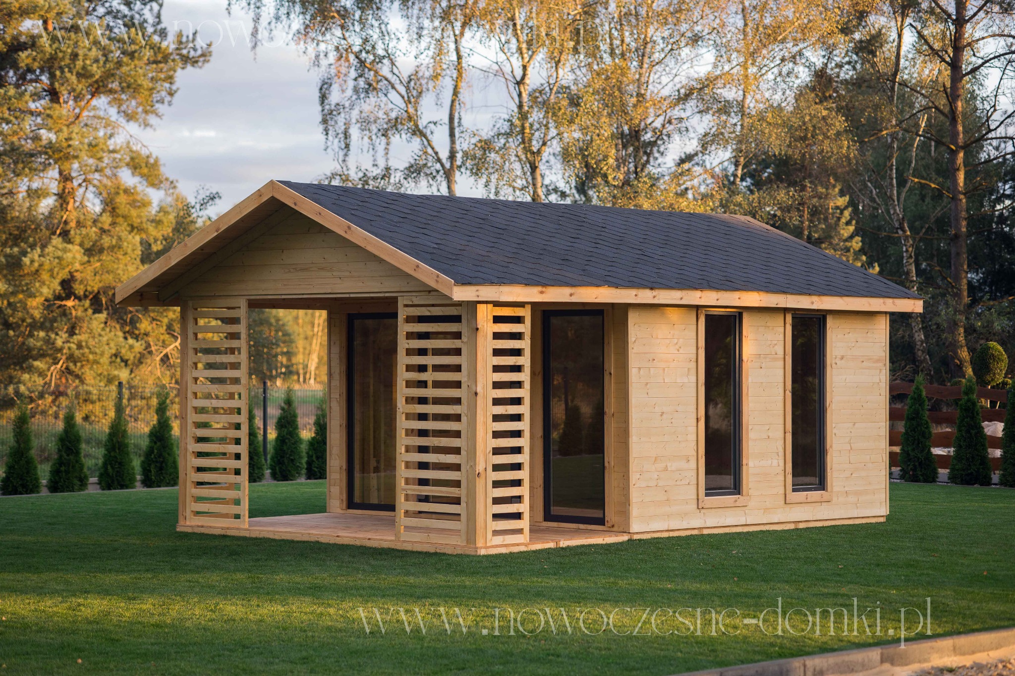 Nowoczesna, drewniana konstrukcja z płaskim dachem i elementami dekoracyjnymi z listew, otoczona zielonym trawnikiem i drzewami.