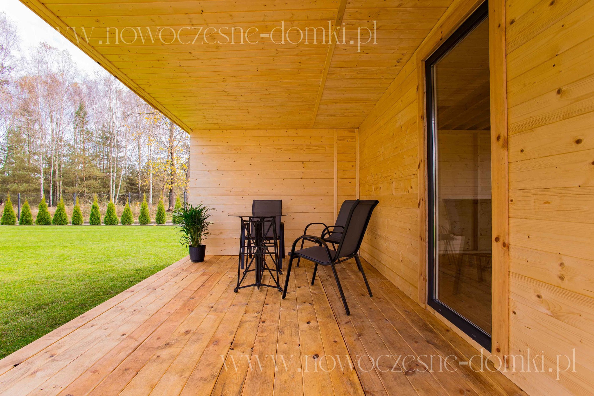 Drewniana konstrukcja z tarasem, na którym stoją czarne krzesła i stolik, widok na trawnik i drzewa w tle.