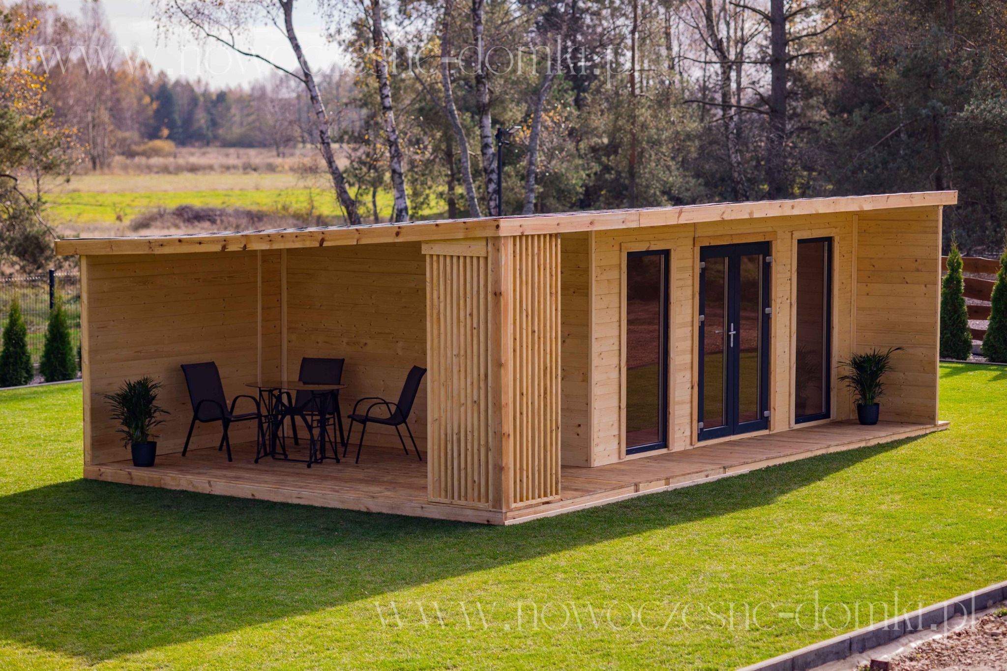 Nowoczesna, drewniana altana z zadaszonym tarasem, wyposażona w czarny zestaw mebli ogrodowych, otoczona zielonym trawnikiem i drzewami w jesiennej scenerii.