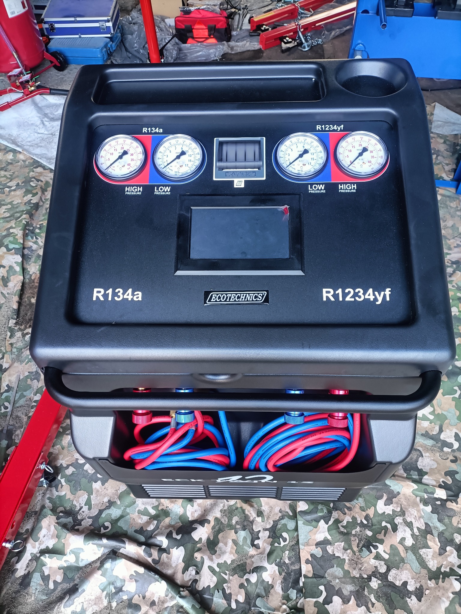 Maszyna do obsługi klimatyzacji samochodowej Ecotecnics, widoczne manometry wskazujące ciśnienie R134a i R1234yf, węże w kolorze czerwonym i niebieskim schowane w dolnej części urządzenia, tło...