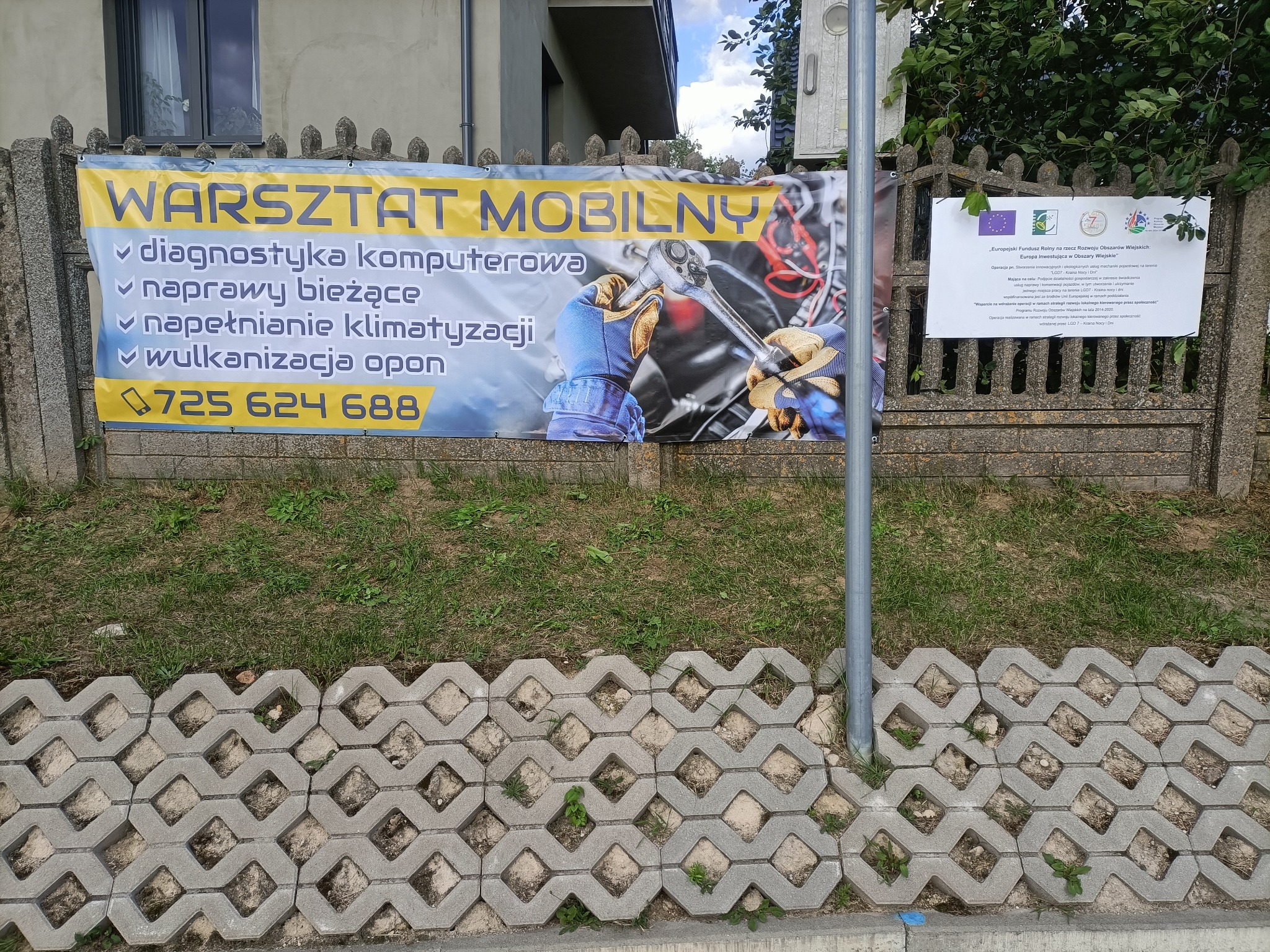 Baner reklamowy mobilnego warsztatu samochodowego z numerem telefonu, diagnostyką komputerową, naprawami bieżącymi, napełnianiem klimatyzacji i wulkanizacją opon, umieszczony na betonowym ogrodzeniu.