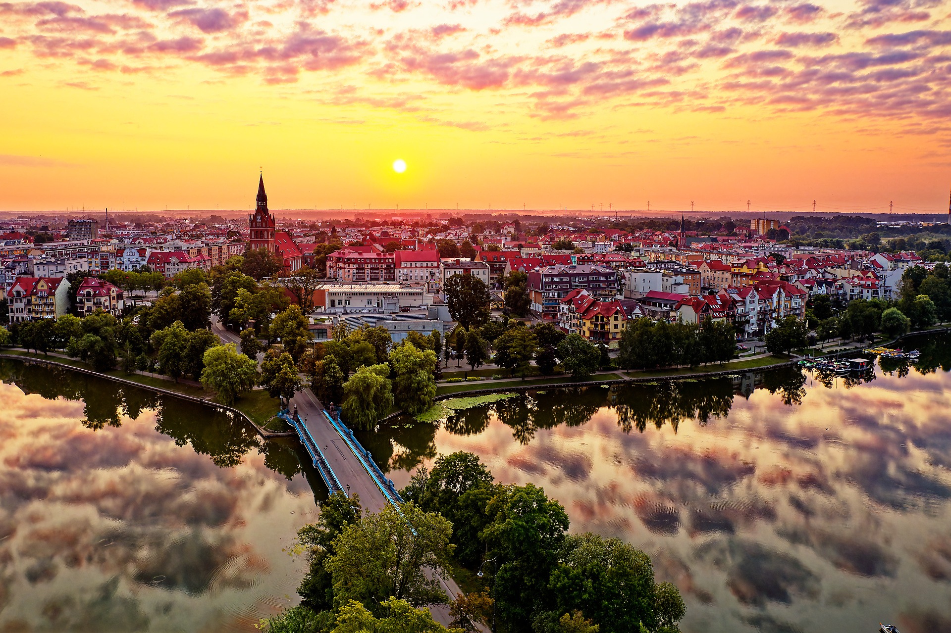 Panorama miasta z lotu ptaka o wschodzie słońca, odbicie chmur w wodzie, most nad rzeką, kościół z wieżą.