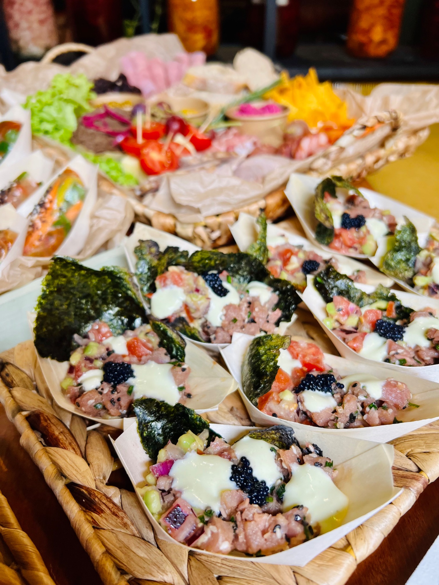 Miniaturowe tarty z surową rybą, warzywami, kawiorem i sosem śmietanowym, podane w biodegradowalnych łódeczkach na plecionym koszu, z półmiskami przekąsek w tle.