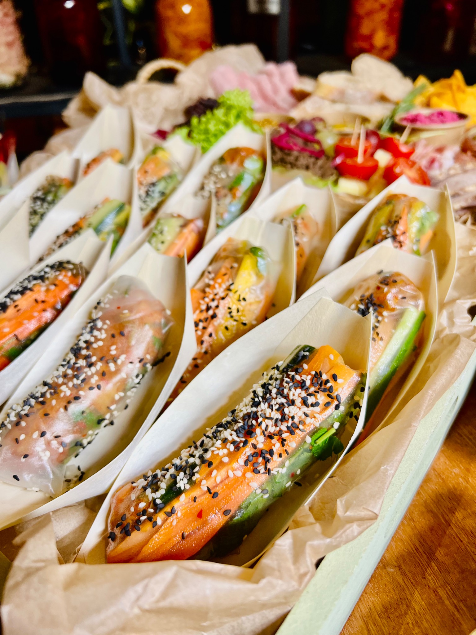 Elegancko podane, transparentne sajgonki z łososiem, ogórkiem i posypką z czarnego i białego sezamu w papierowych łódeczkach, na tle bufetu z przekąskami.
