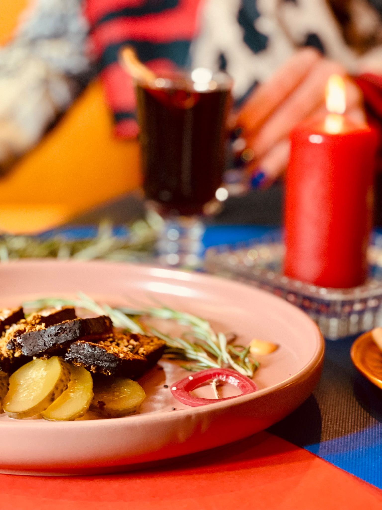 Świąteczne danie z ciemnego pieczywa z posypką, ogórkami kiszonymi i czerwoną cebulą na różowym talerzu, w tle grzane wino z pomarańczą i płonąca czerwona świeca, tworzące kameralną atmosferę.