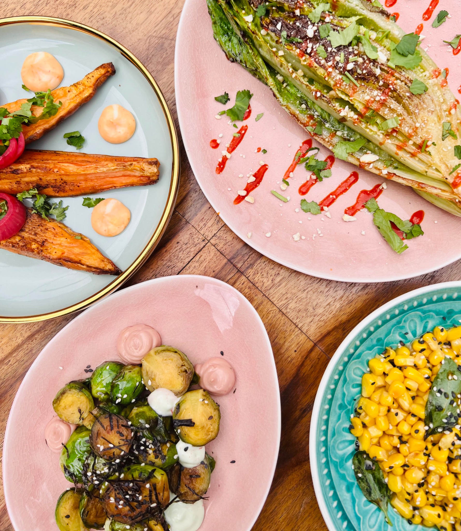 Widok z góry na cztery talerze z różnymi daniami: pieczone bataty z sosem i cebulą, grillowana sałata rzymska z sosem i ziołami, brukselka z sosem i sezamem oraz kukurydza z sosem i sezamem...